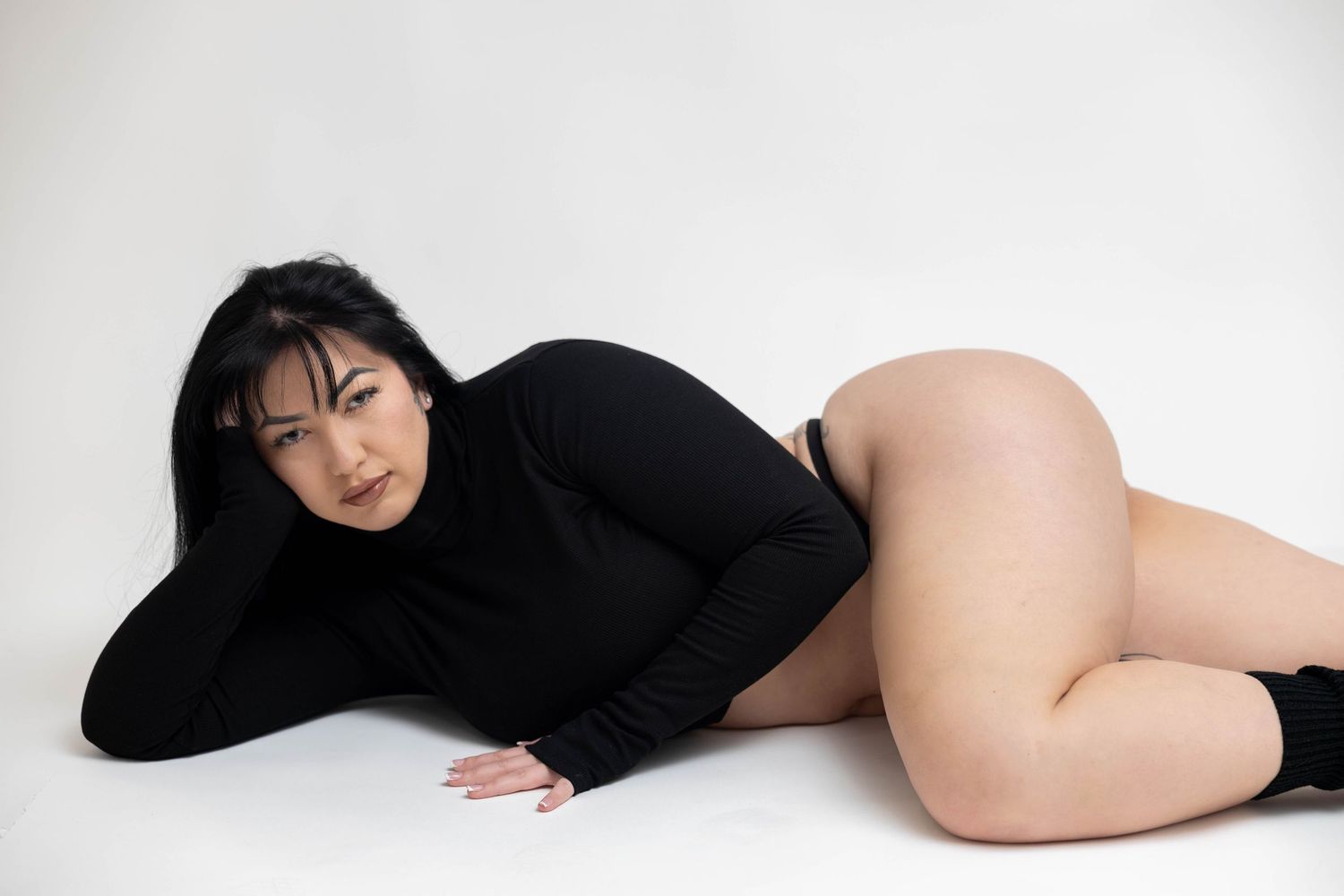 A woman in a black long sleeve bodysuit poses gracefully in a minimalist studio setting.