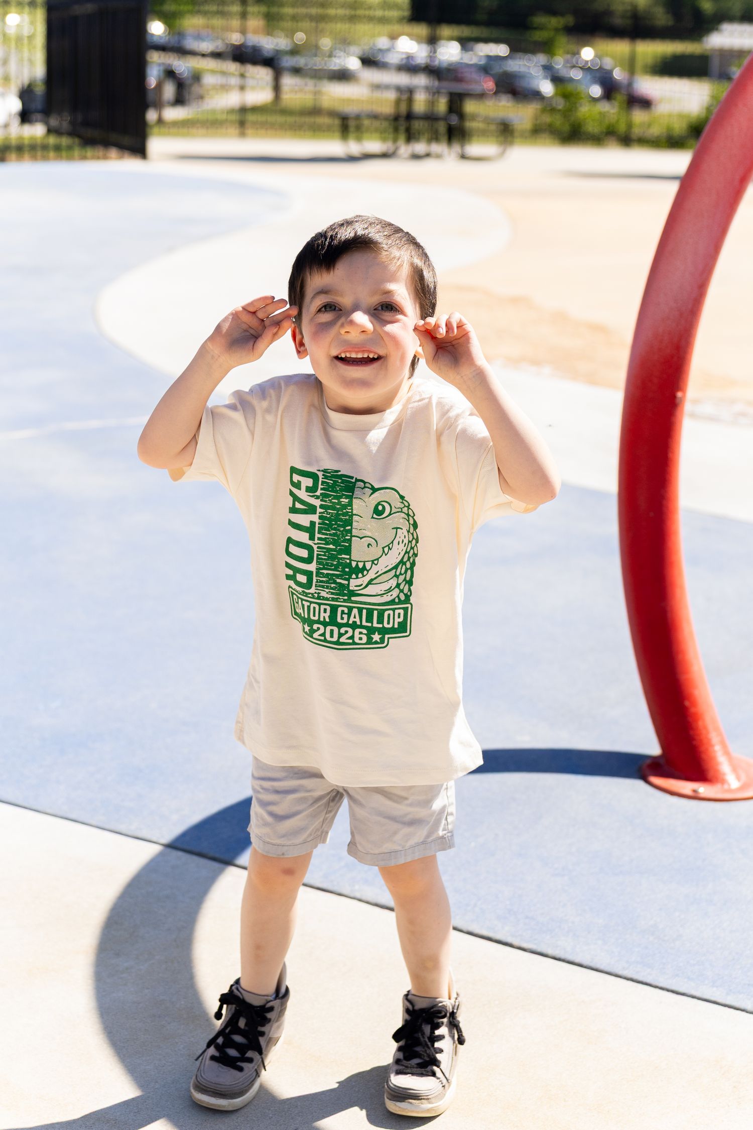 Smiling young boy wearing a Gator Gallop 2026 t-shirt and gray shorts, playing at an outdoor playground.