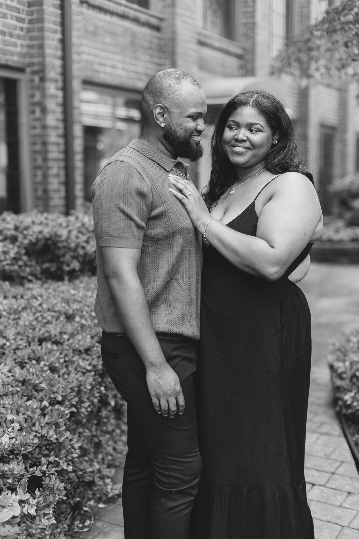 Black and white photo of a smiling couple embracing outdoors near brick building and lush greenery.