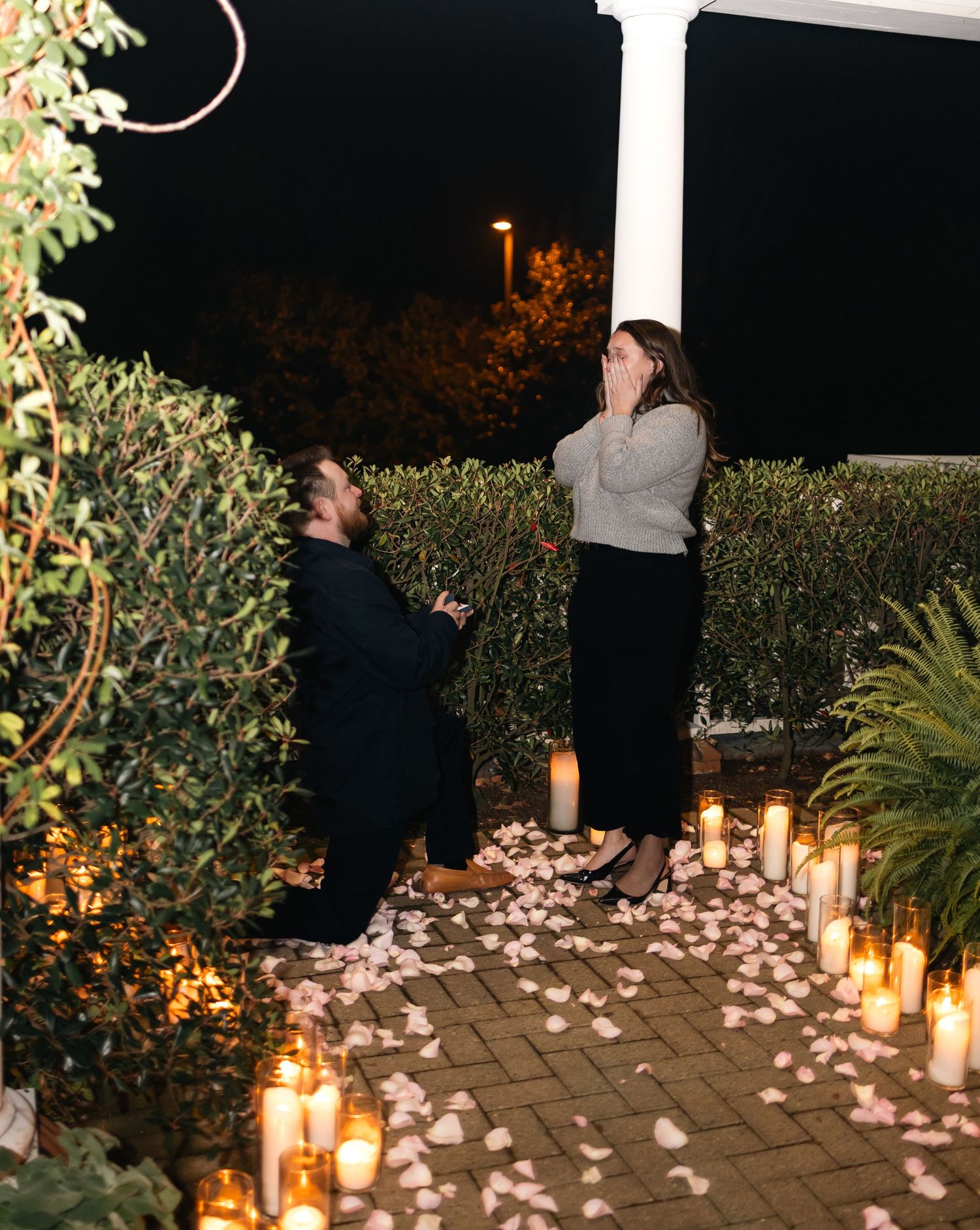 A romantic outdoor proposal setup with glowing candles and scattered rose petals on a paved walkway at night.