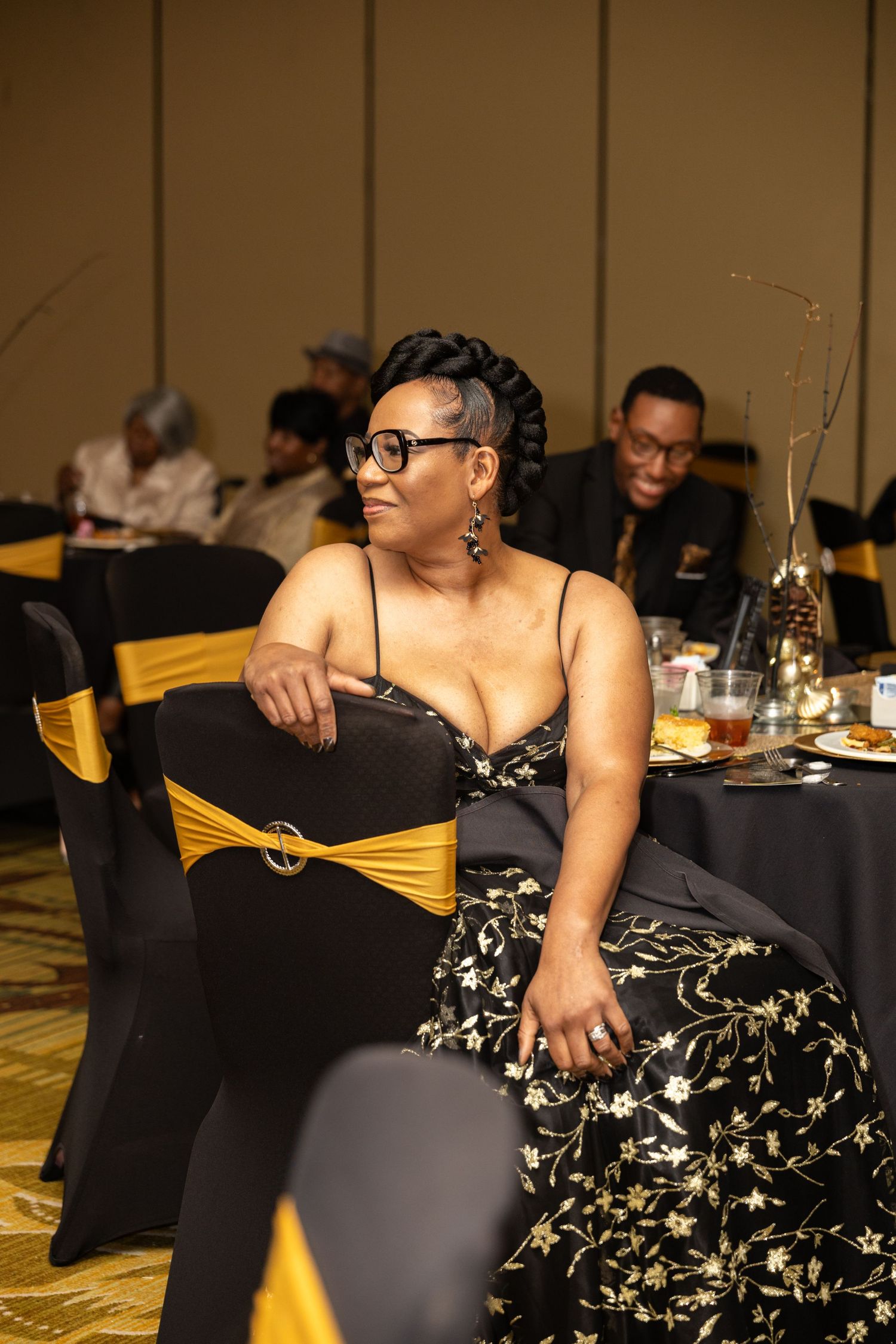 A person in a black and white floral dress sits at a formal event table with dark tablecloths.