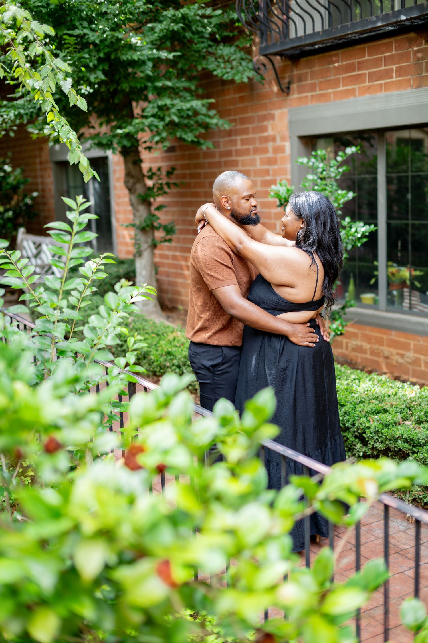 A couple embraces romantically in a lush garden courtyard beside a brick building, the woman in a black dress.