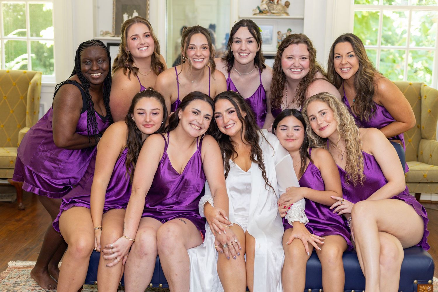 Bride in white robe poses with bridesmaids in purple satin dresses during wedding preparations.