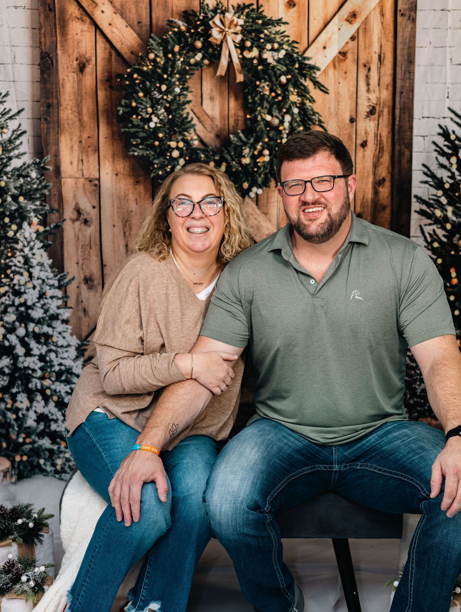 Couple sitting on wooden bench with winter decor including wreaths and flocked trees against rustic wood backdrop.