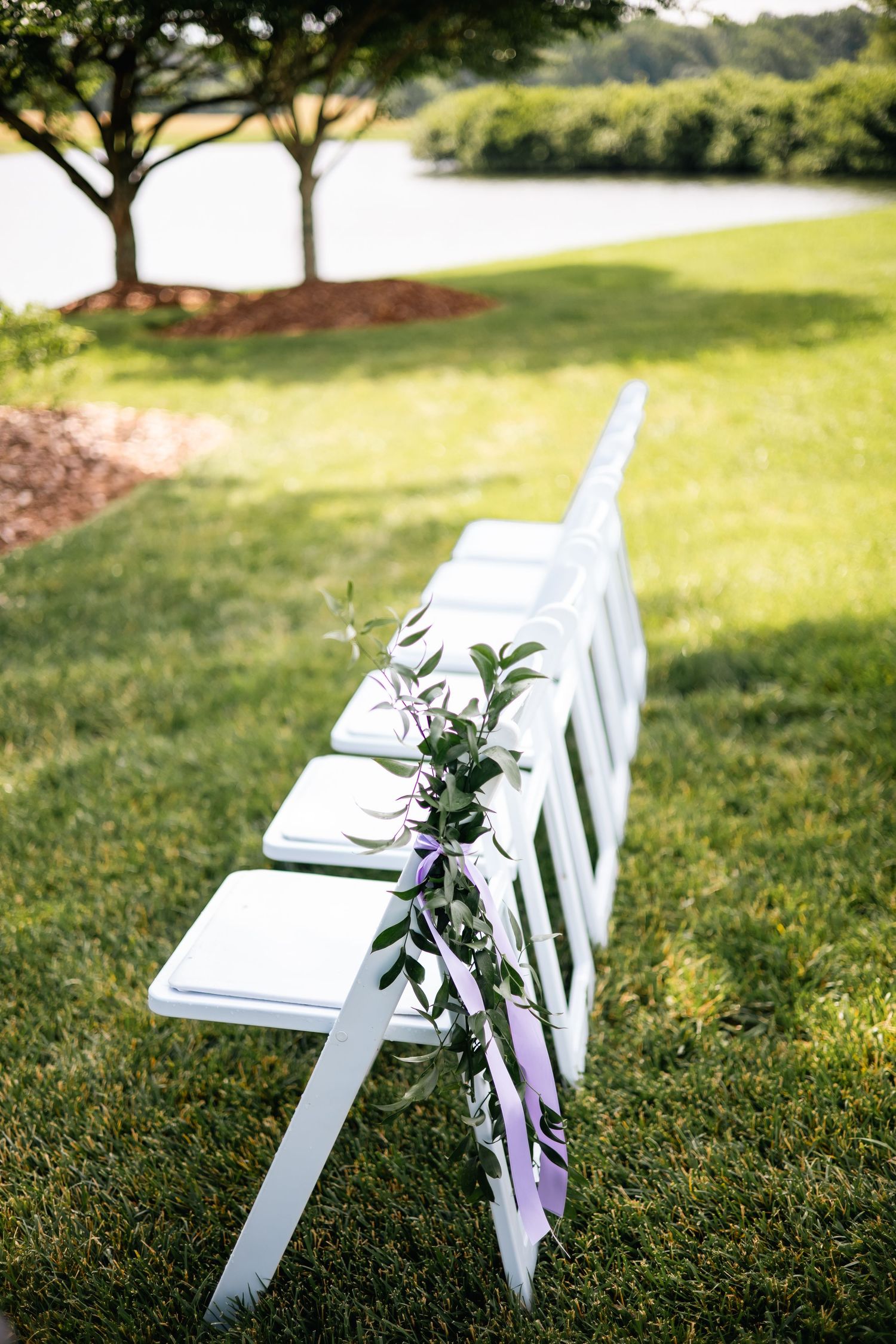 White folding chairs decorated with purple ribbons and floral arrangements set up on a grassy lawn near a waterfront.