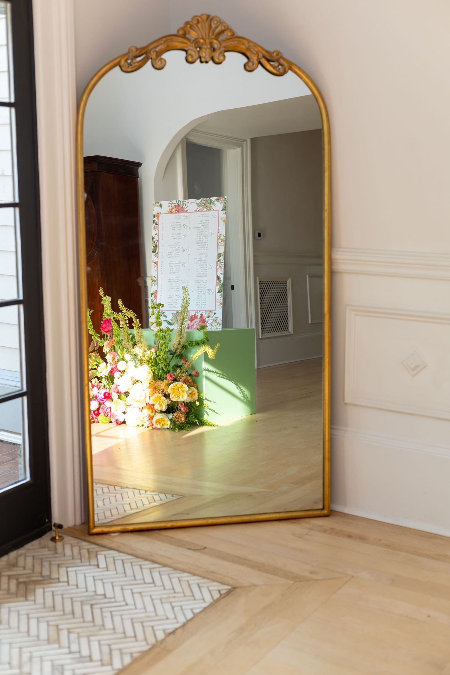 Ornate gold arched floor mirror reflecting colorful floral arrangements in a bright, elegant room with herringbone floors.