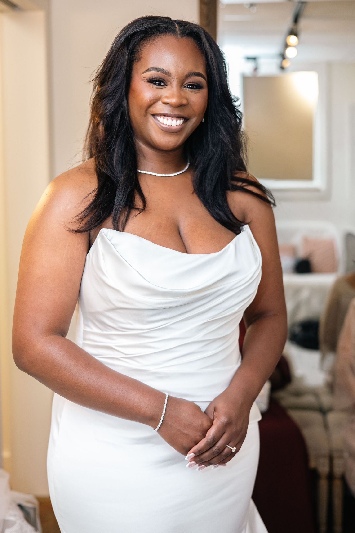 A woman in an elegant white dress with a draped neckline and pearl necklace smiles brightly in an indoor setting.