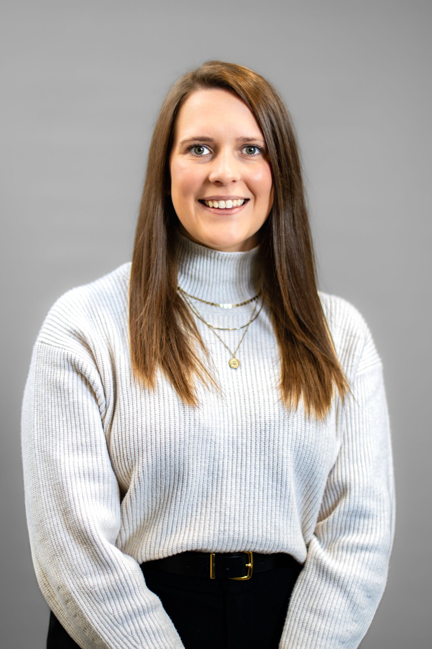 Smiling woman with long brown hair wearing a light gray turtleneck sweater and gold necklaces against a gray background.