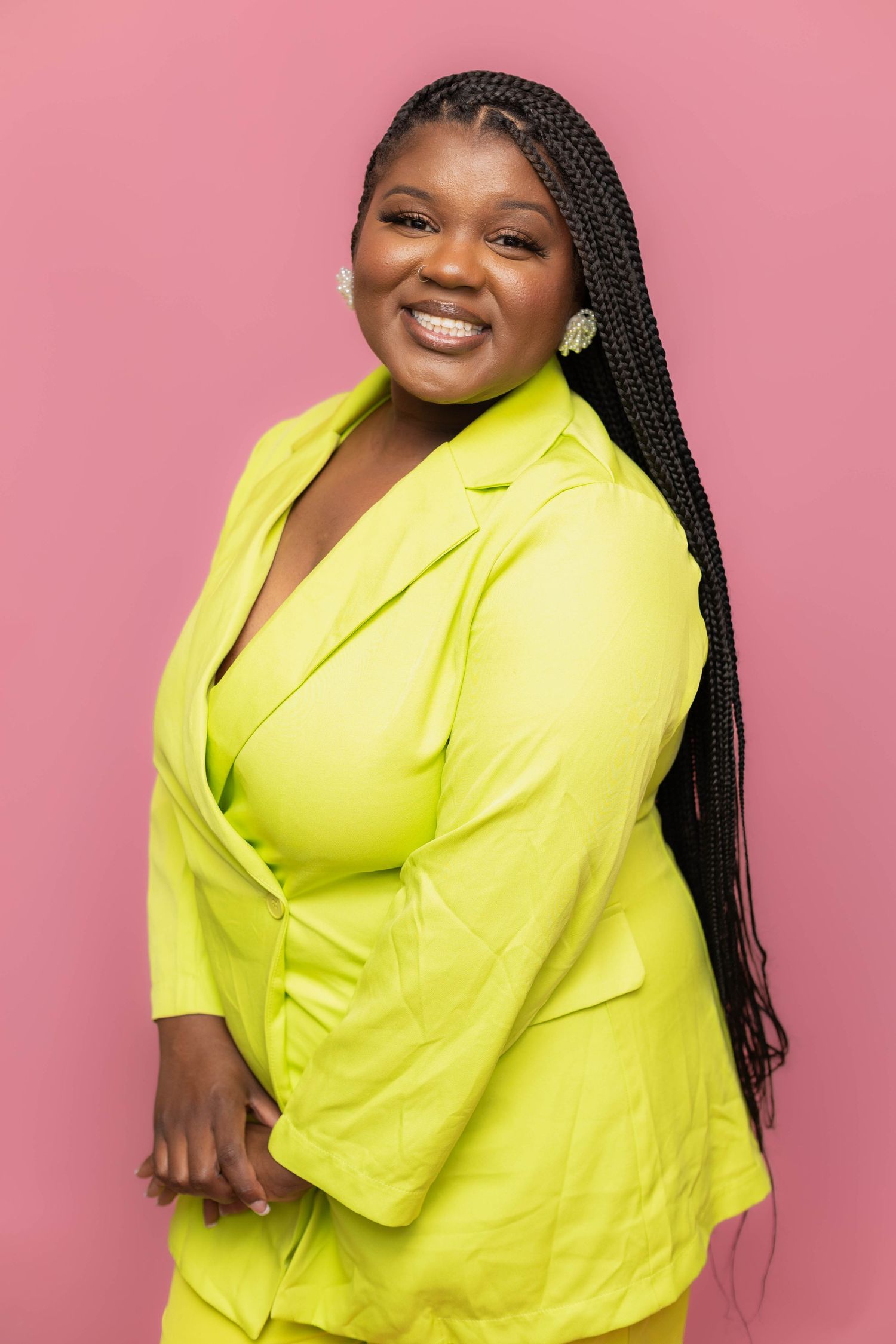 Smiling woman in neon yellow blazer suit with long braids poses confidently against a pink background.