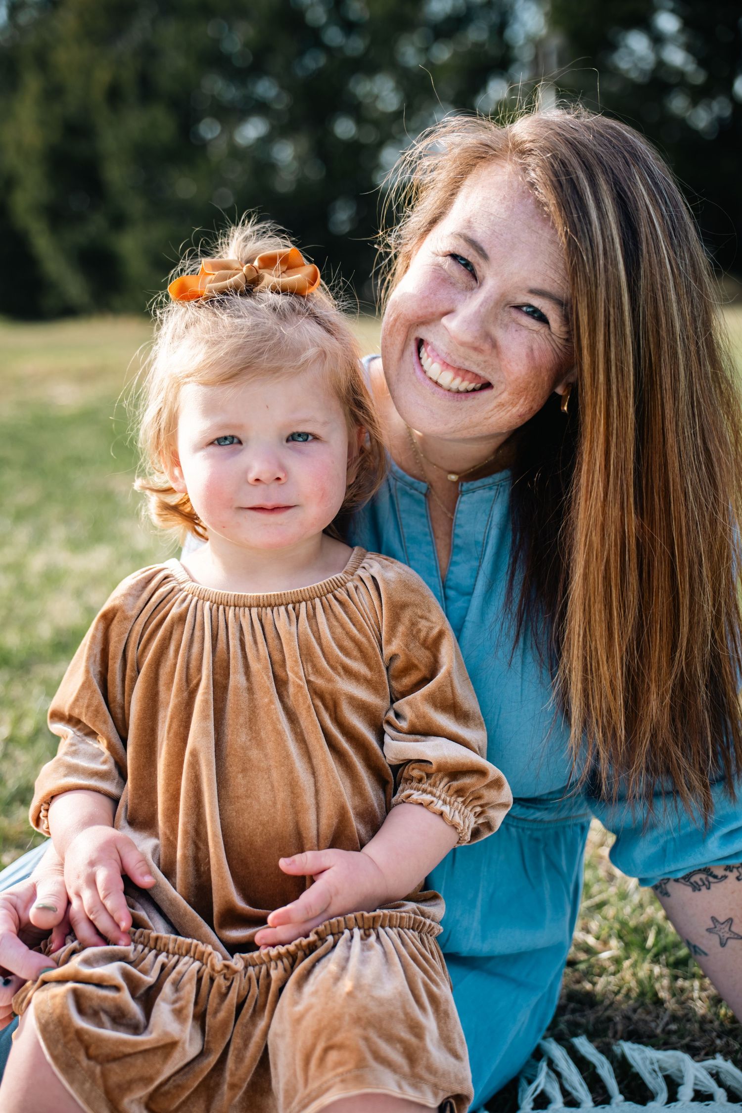 A smiling woman in teal dress poses outdoors with a toddler girl in brown velvet dress wearing an orange bow.