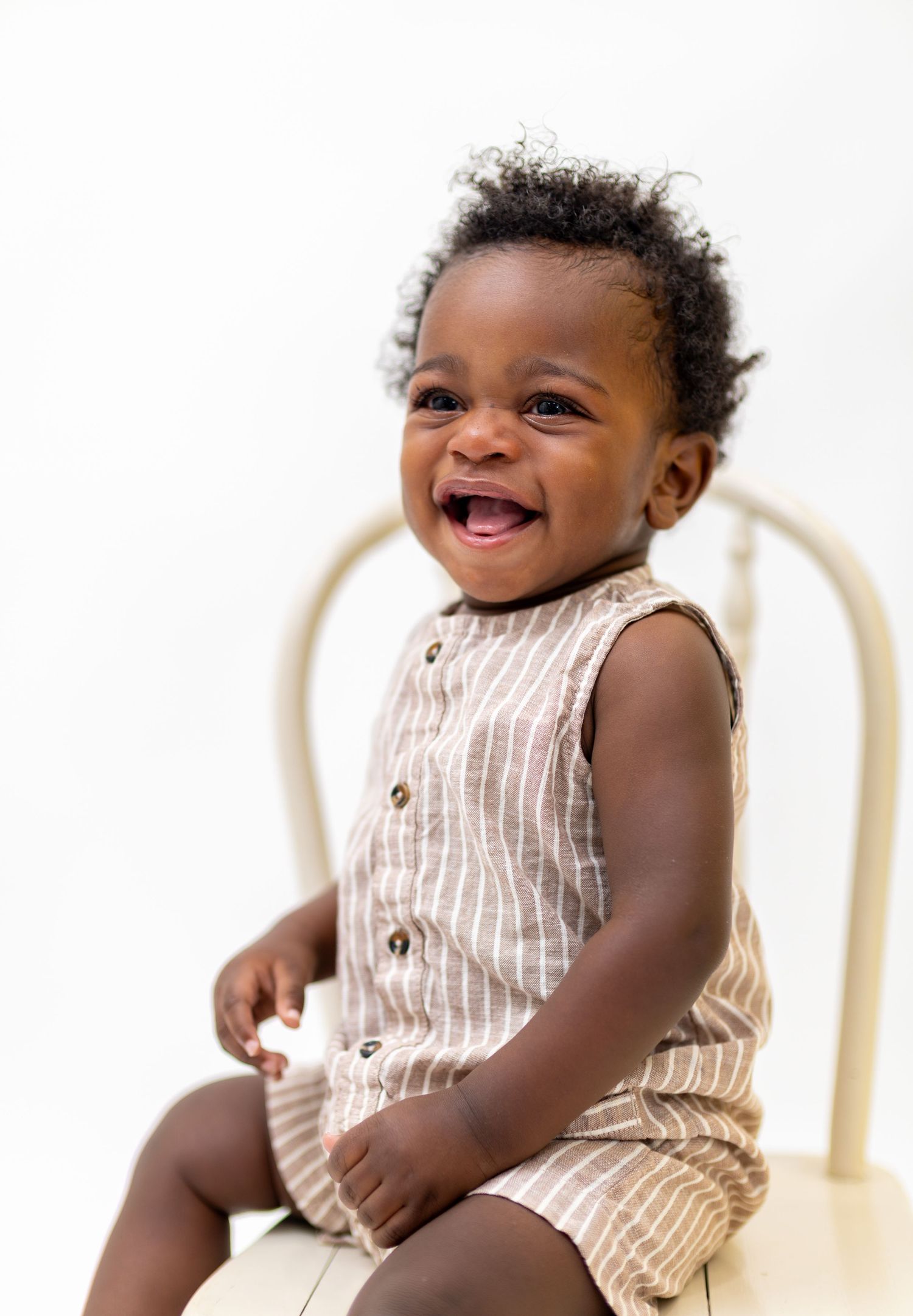 Smiling Black baby wearing a striped linen romper sitting on a white chair against a white background.