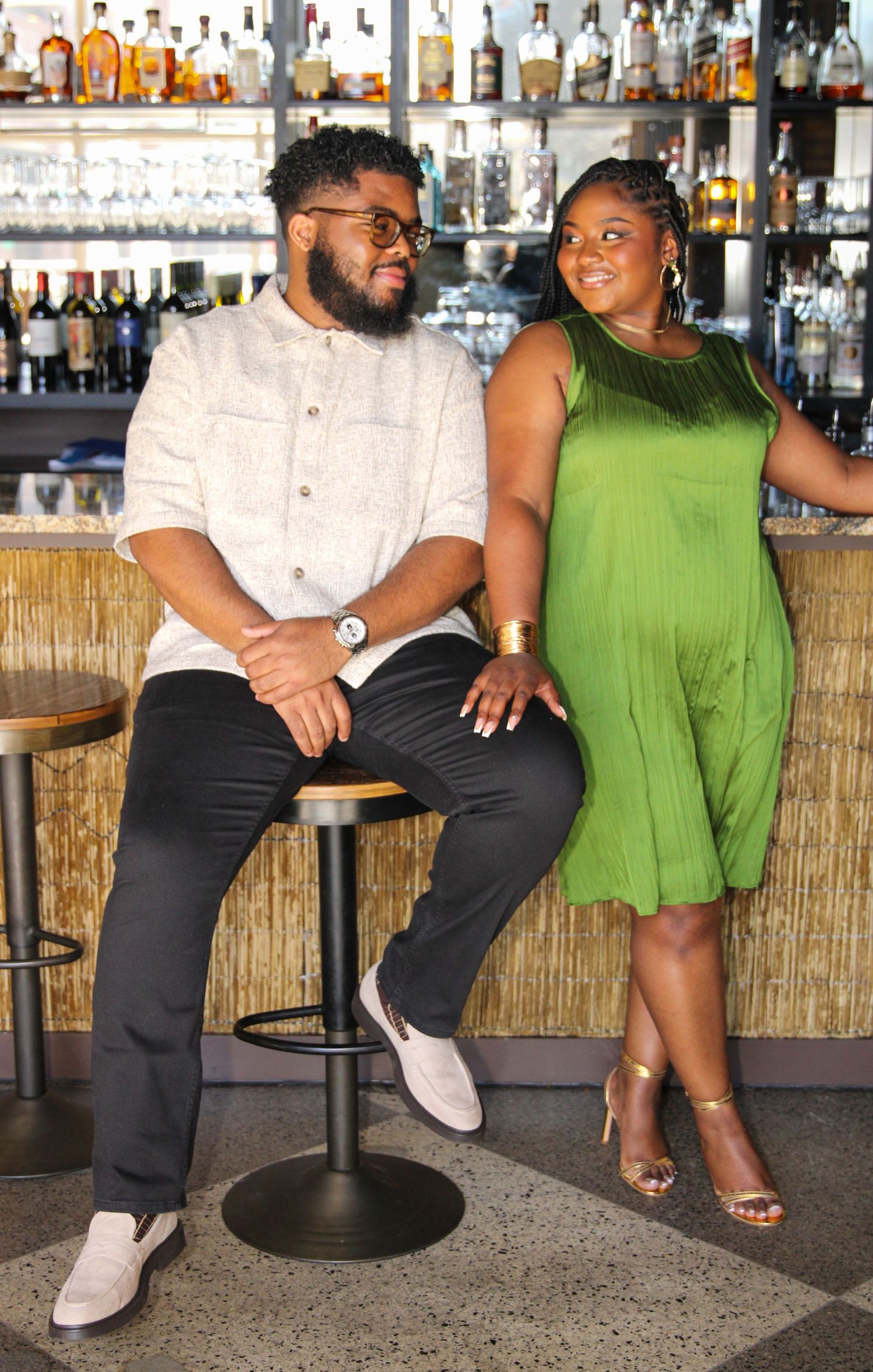 A couple poses together at a bar counter with liquor bottles displayed on shelves behind them.
