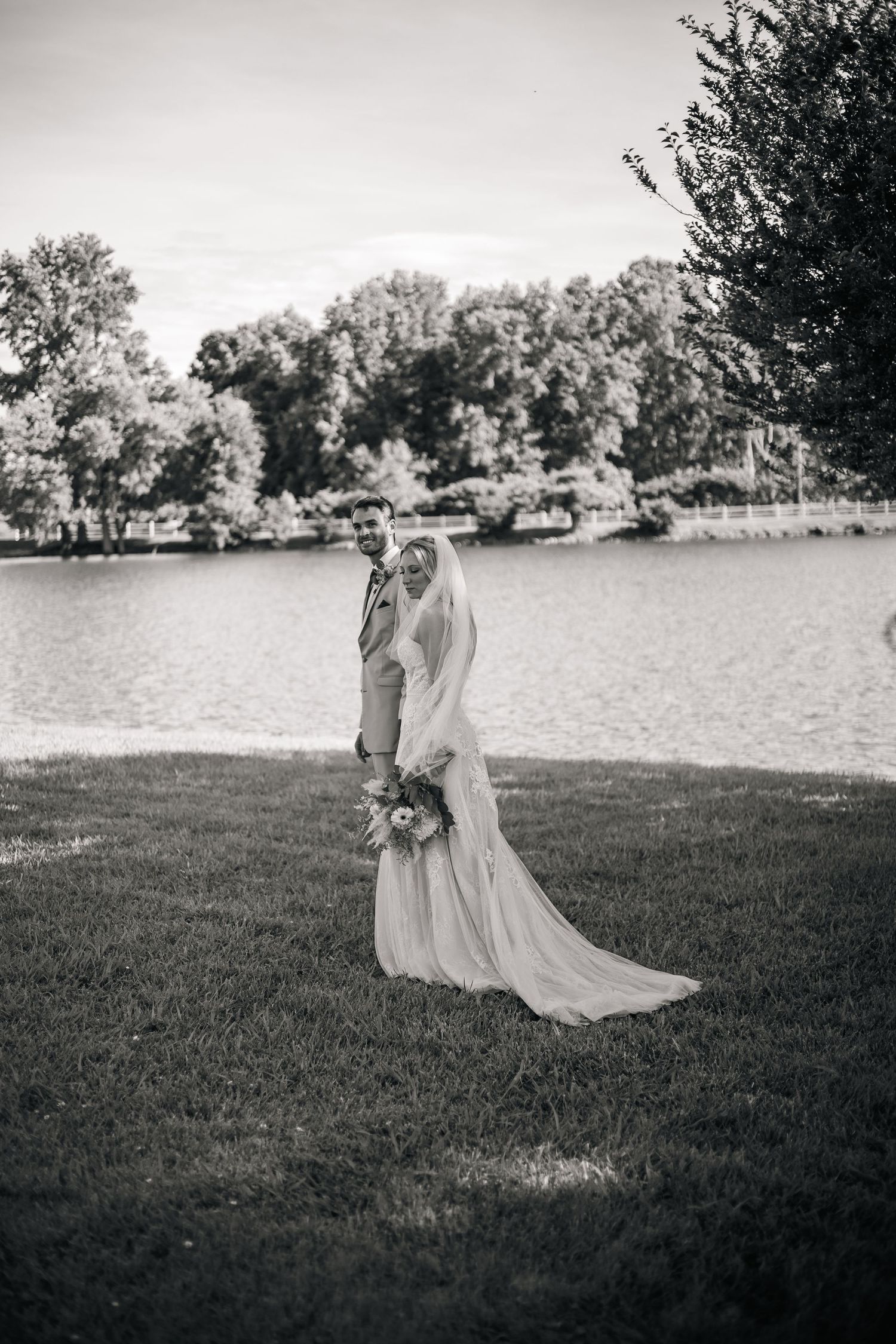 A romantic couple embraces on a grassy lawn by a serene lake surrounded by trees in black and white photography.