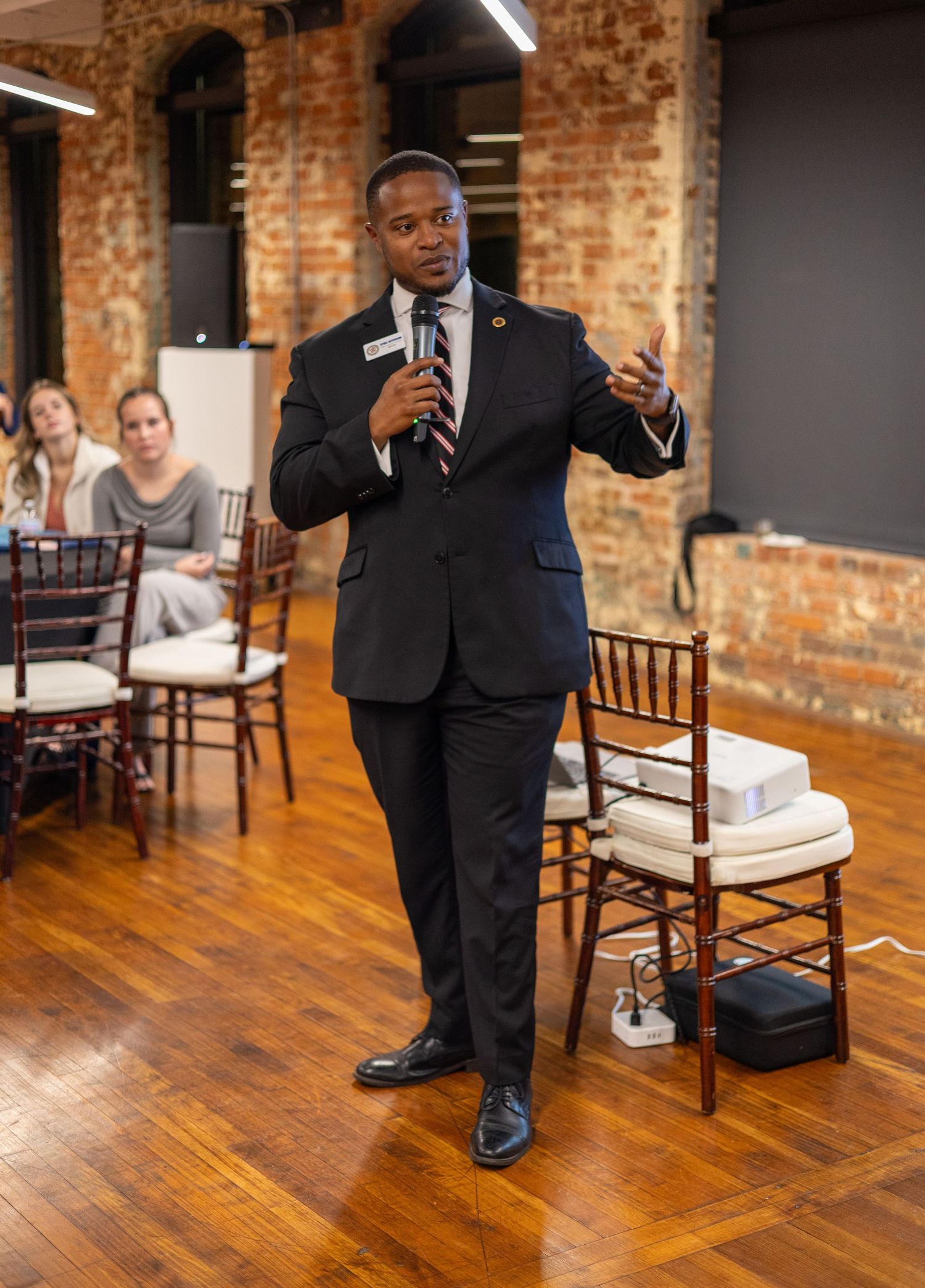 A man in a dark suit speaks into a microphone at an indoor event with brick walls and wooden floors.