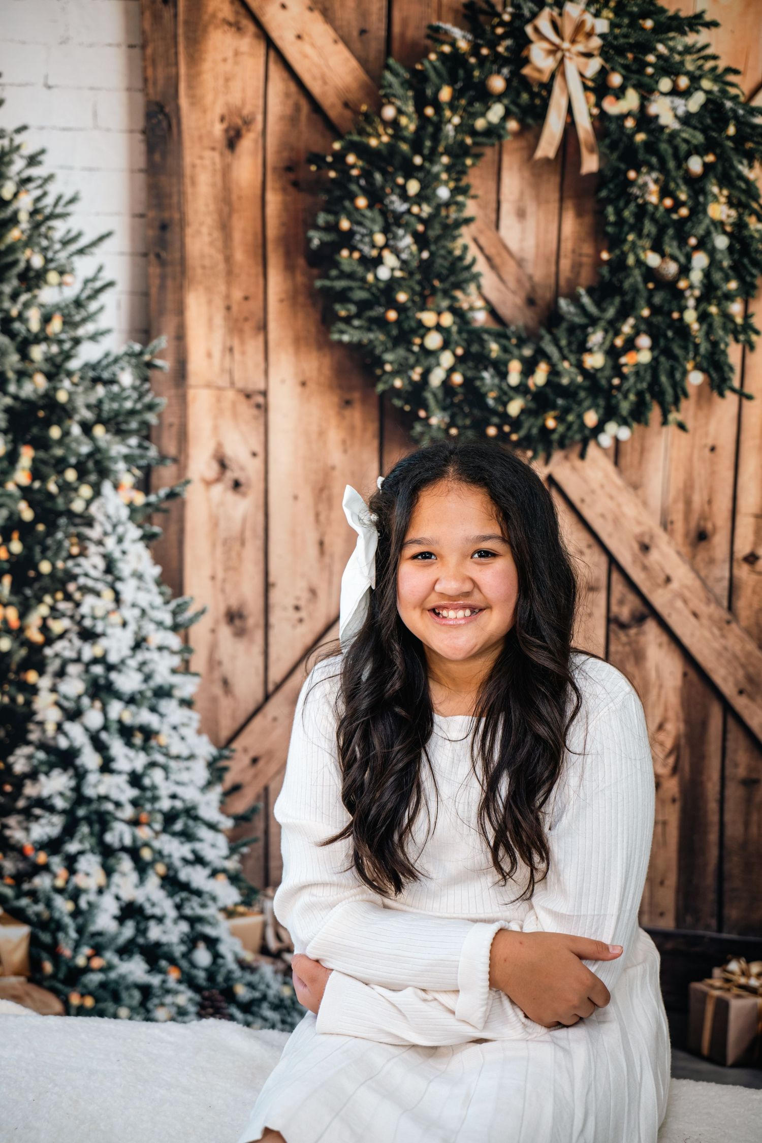 A festive winter scene with a wooden barn door backdrop decorated with a large wreath and snow-dusted Christmas tree.
