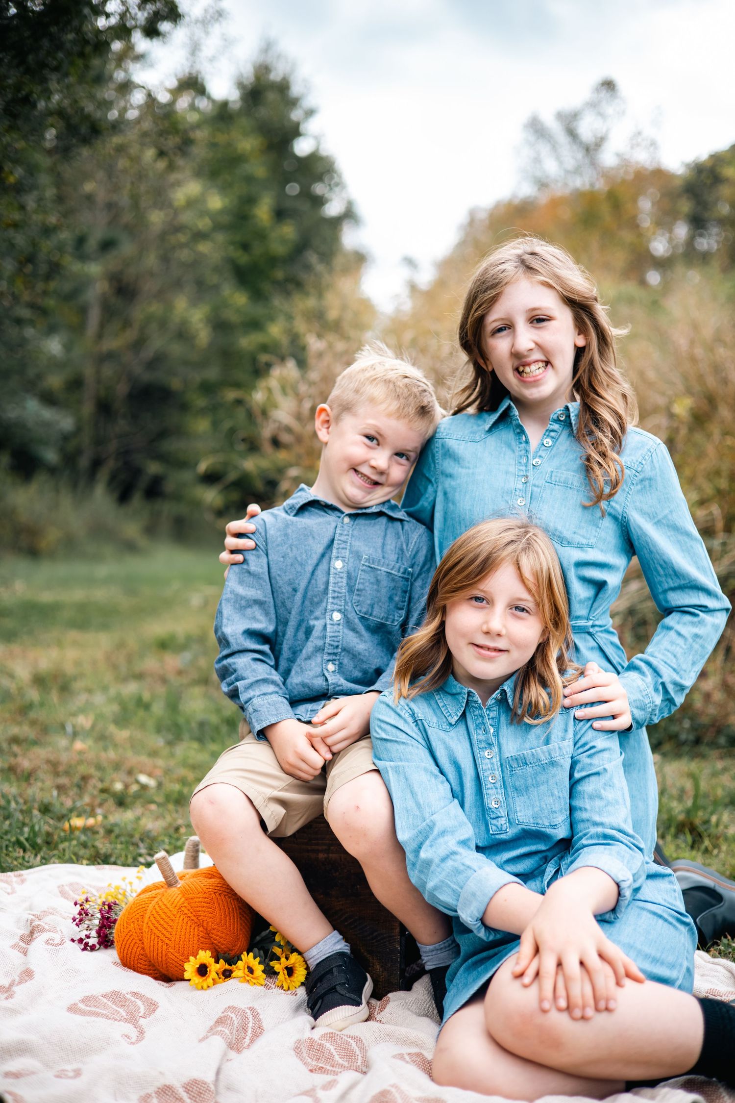 Family wearing coordinating denim outfits poses on blanket with pumpkin and sunflowers during autumn outdoor photo session.