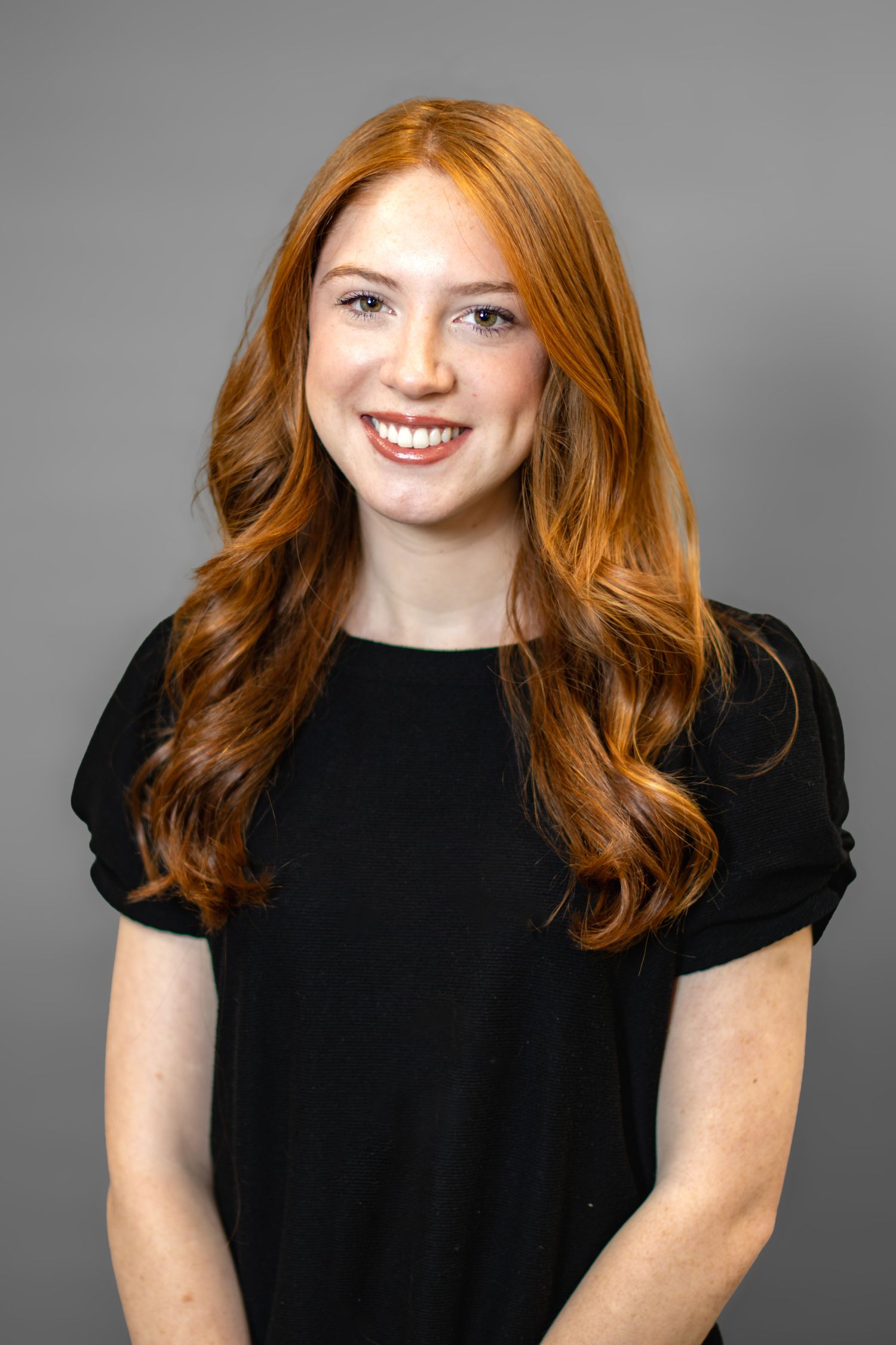 Young woman with long red hair smiling, wearing a black top against a gray background.