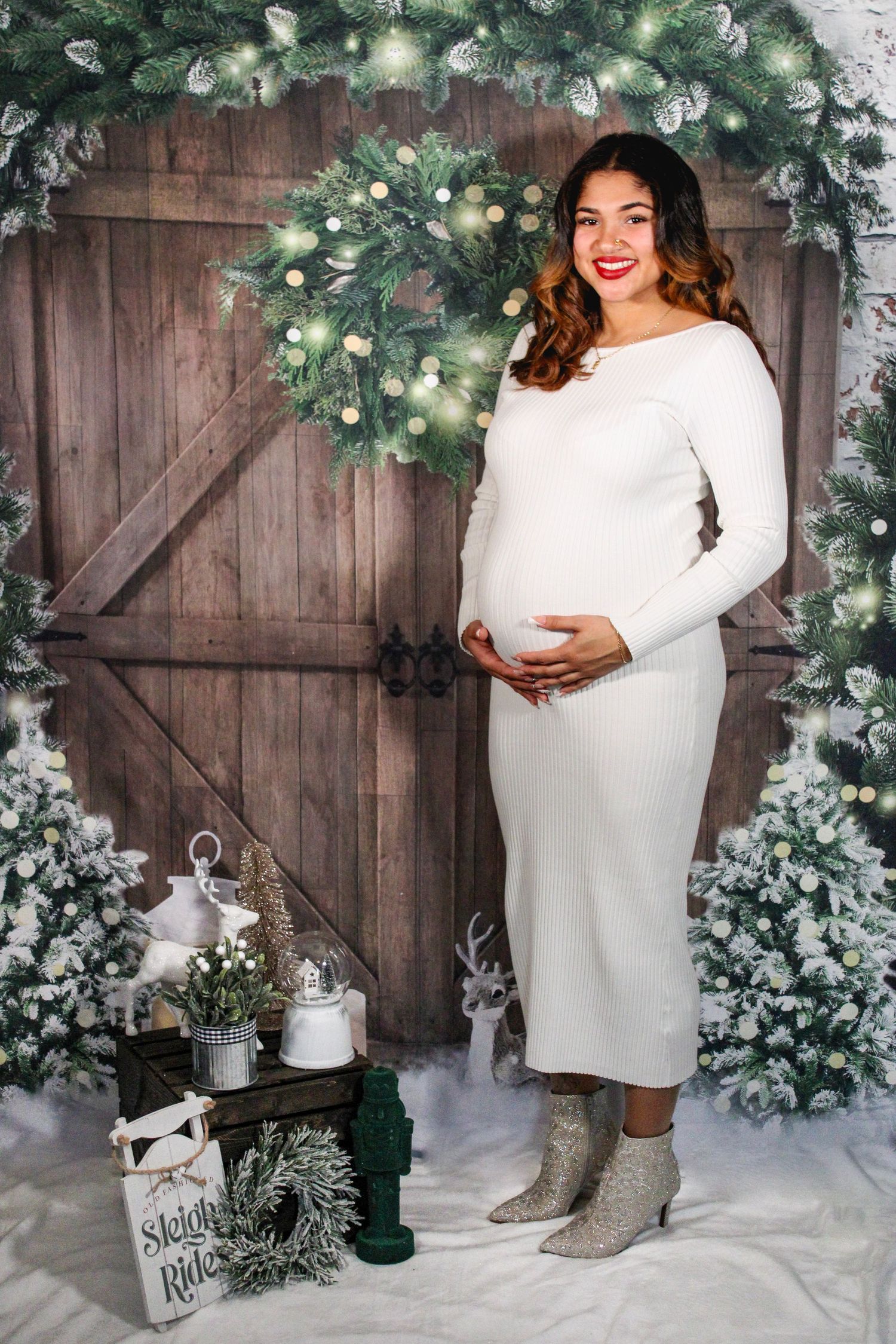 Maternity photo of person in white dress against rustic barn door with holiday wreath and decorations.