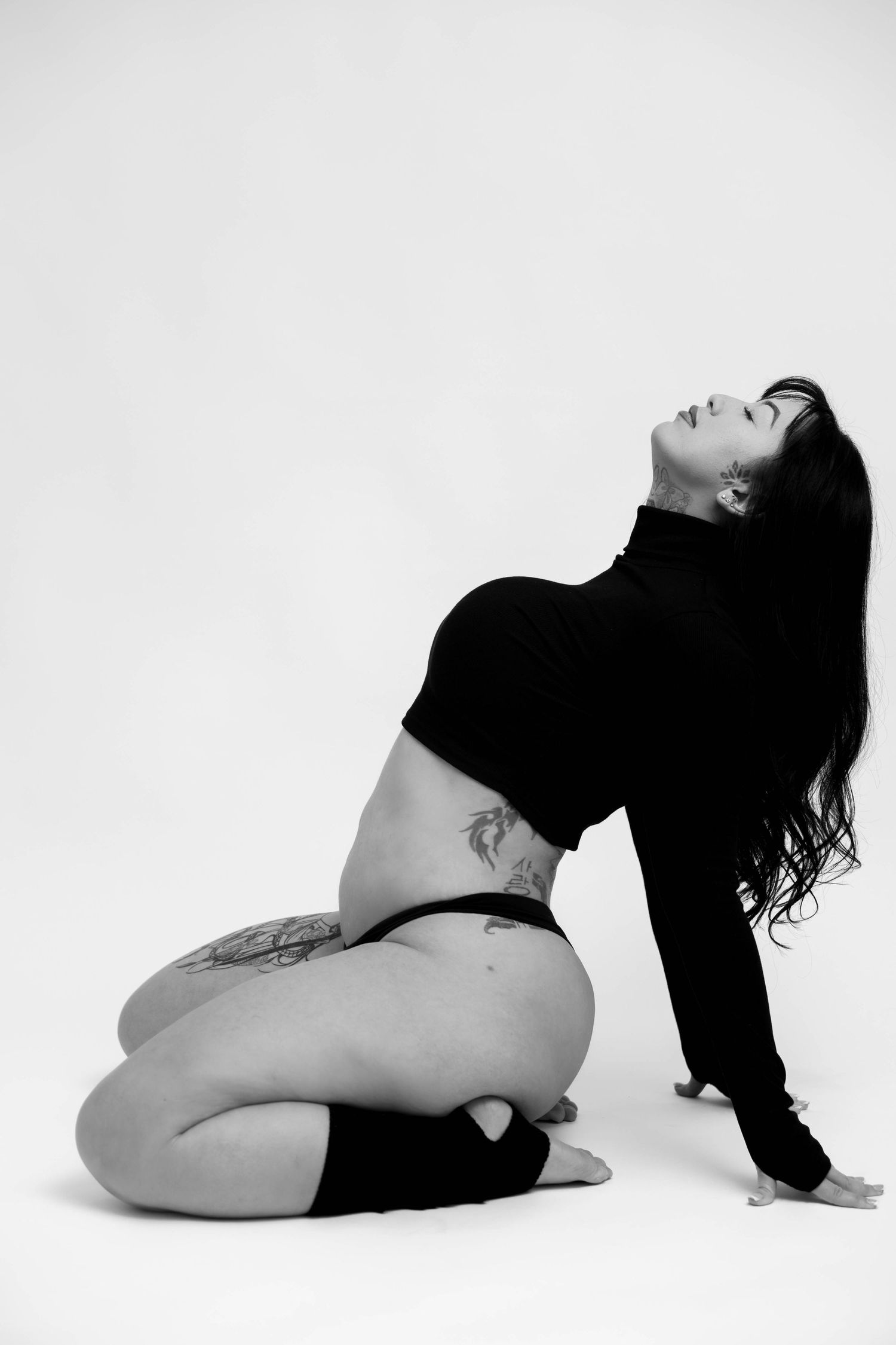 Black and white artistic portrait of a model in a cropped top showing tattoos in a dramatic pose against white backdrop.
