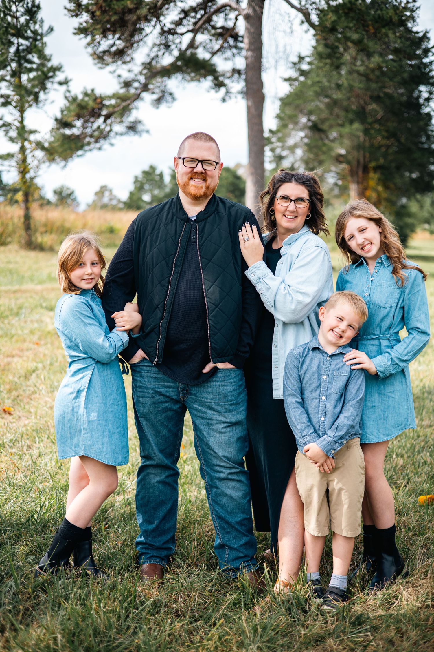 A family of five poses together outdoors in coordinating denim and blue outfits against a backdrop of tall pine trees.