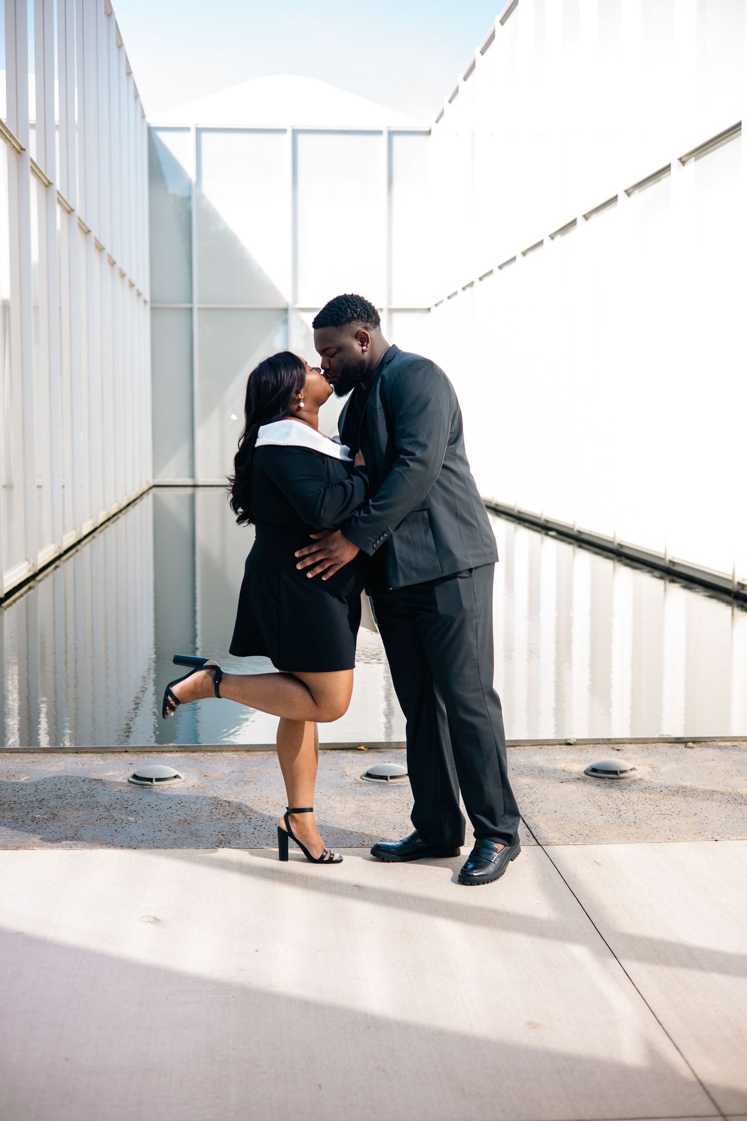 A romantic couple in formal business attire share an intimate moment in a modern hallway with white walls.