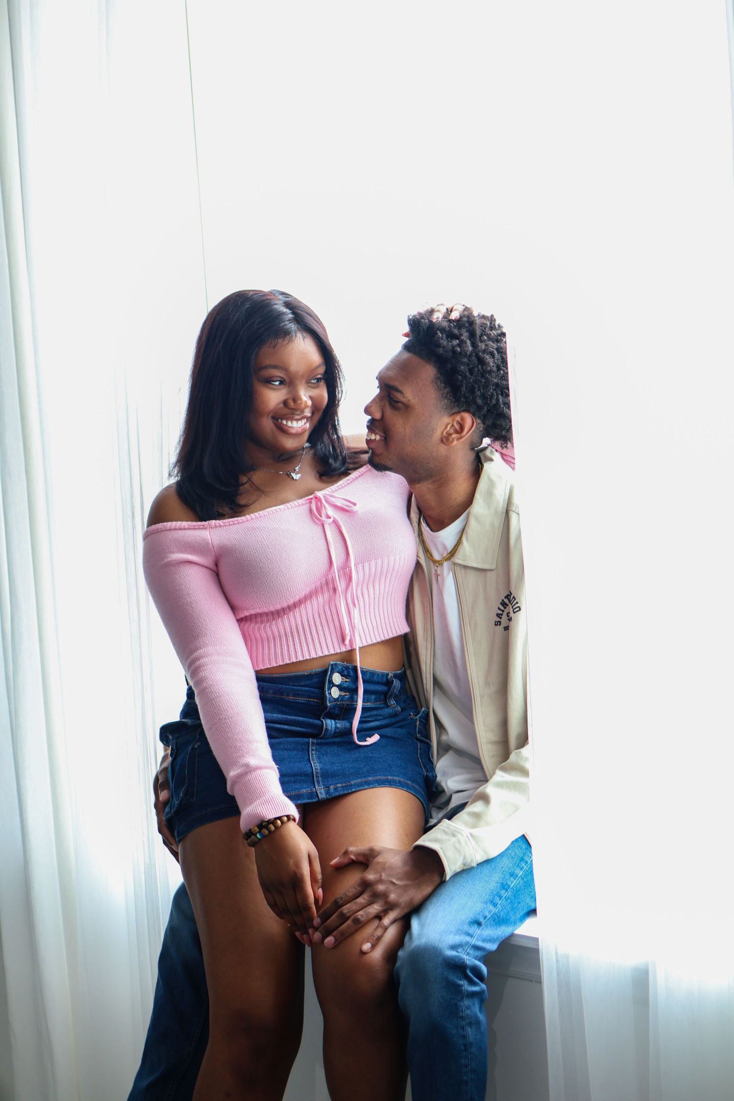 A couple shares an intimate moment by a window, with one wearing a pink top and the other in beige.