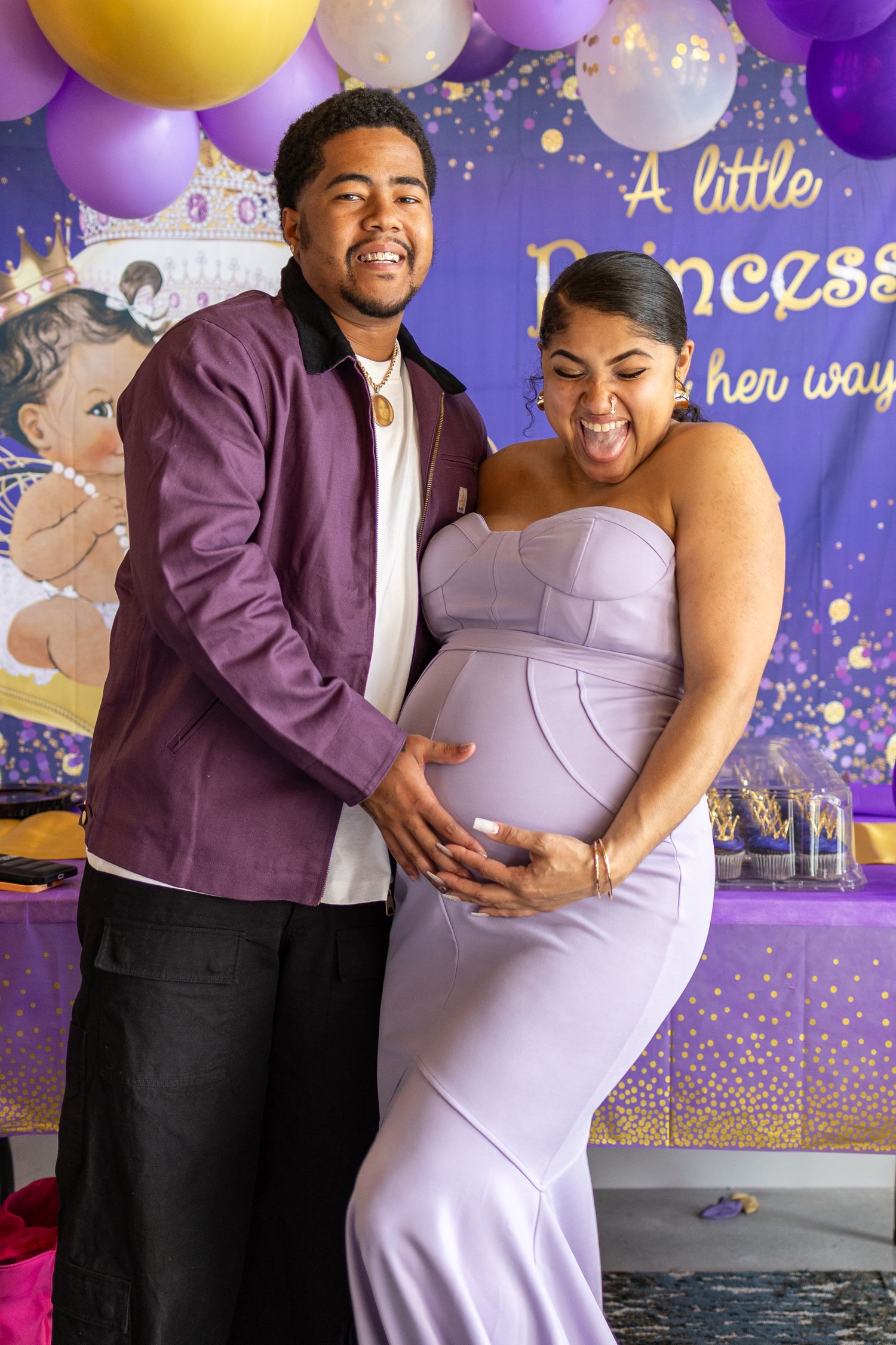 Joyful couple hugging and celebrating at a colorful baby shower with purple and gold balloons and decorations.