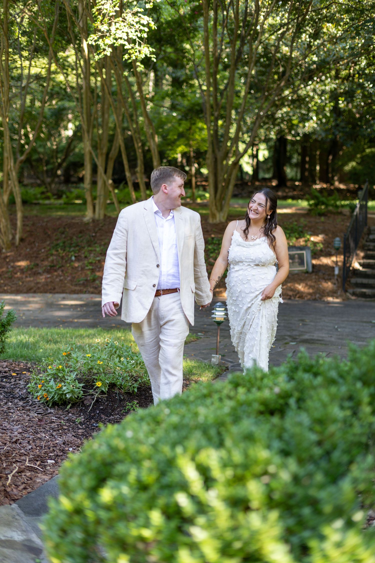 A bride and groom walk hand-in-hand along a garden path surrounded by lush greenery on their wedding day.