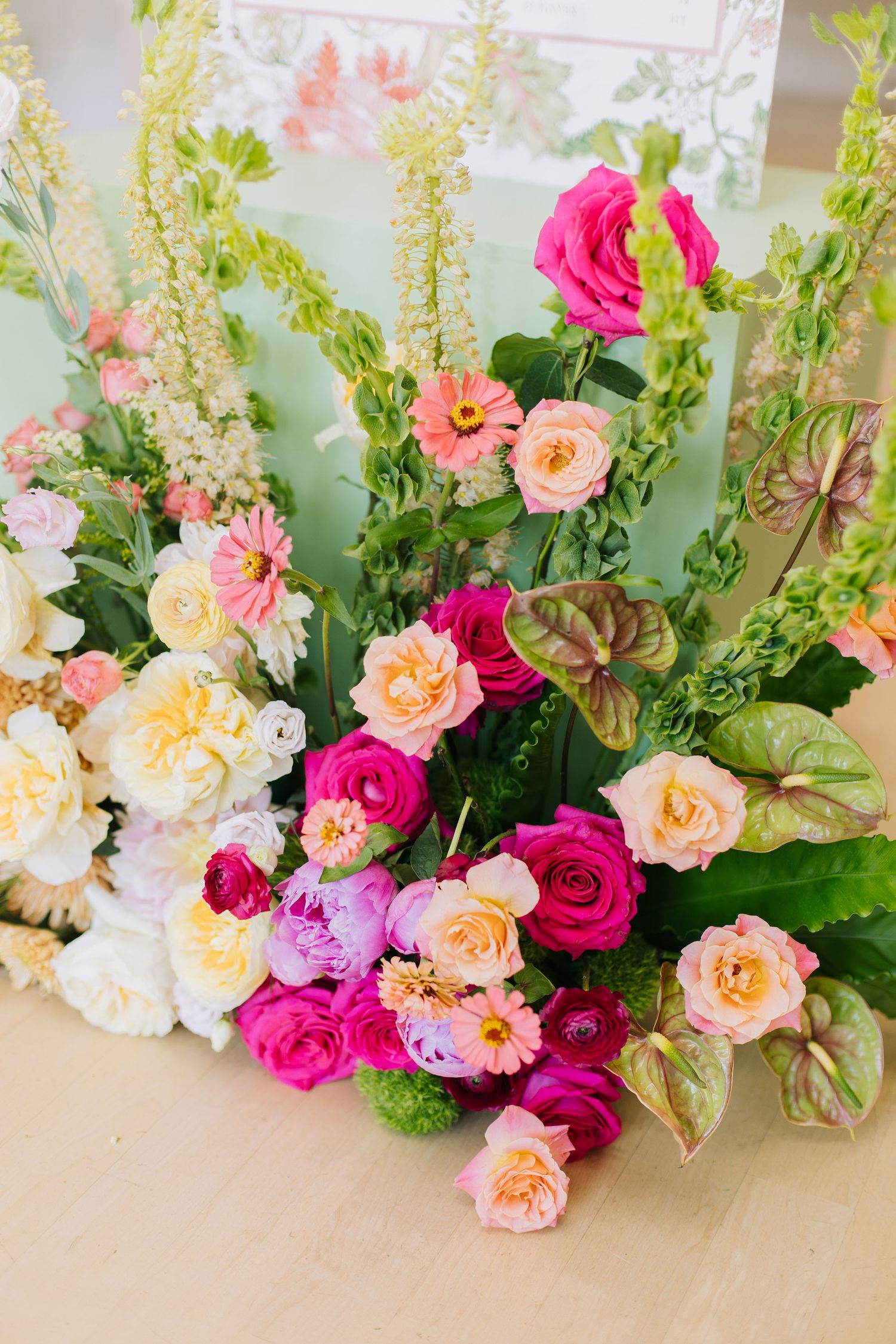 Lush floral arrangement featuring pink roses, magenta blooms, peach ranunculus, and white flowers with green foliage on a table.