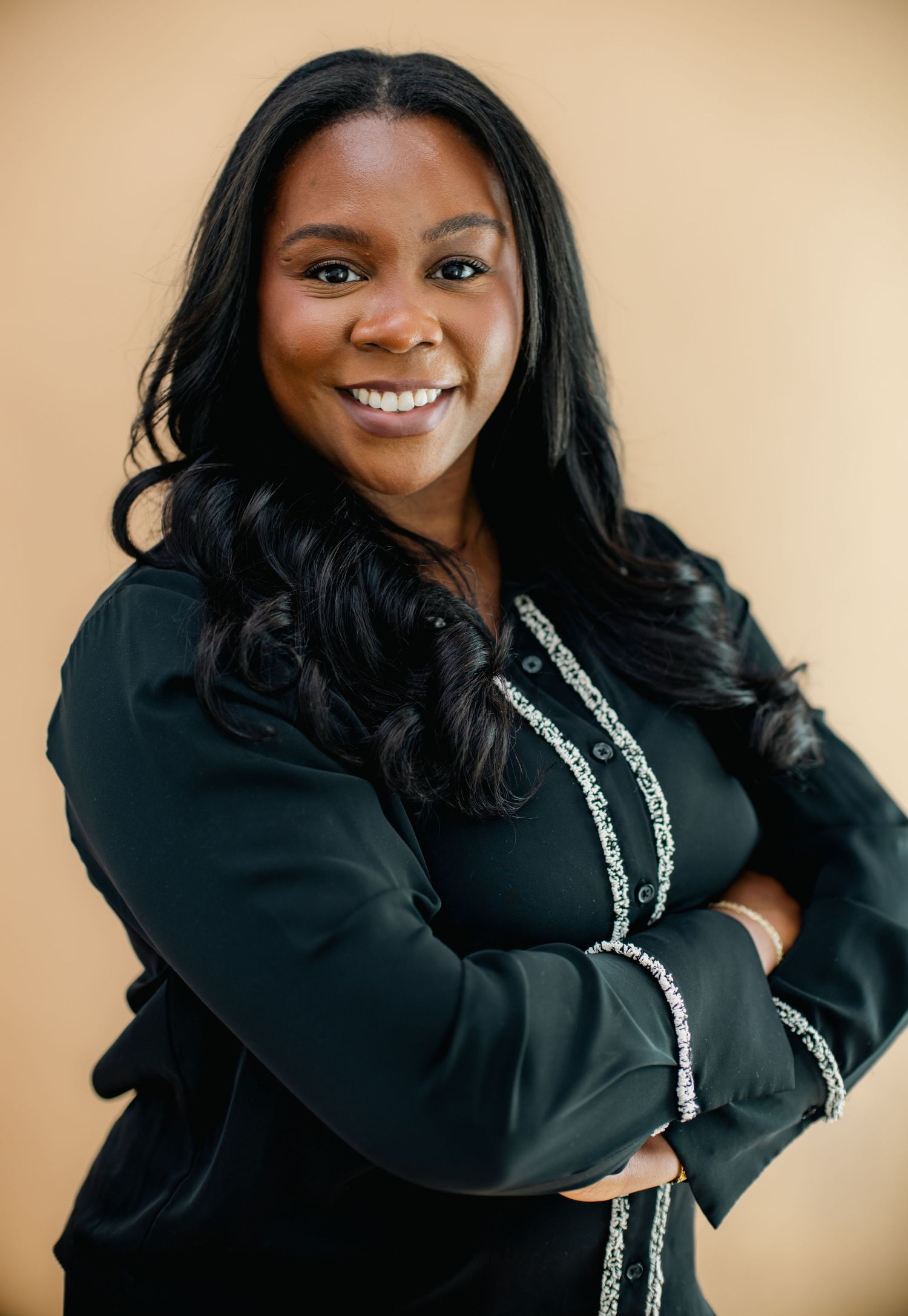 Professional woman in black blazer with arms crossed, smiling confidently against a warm beige background.