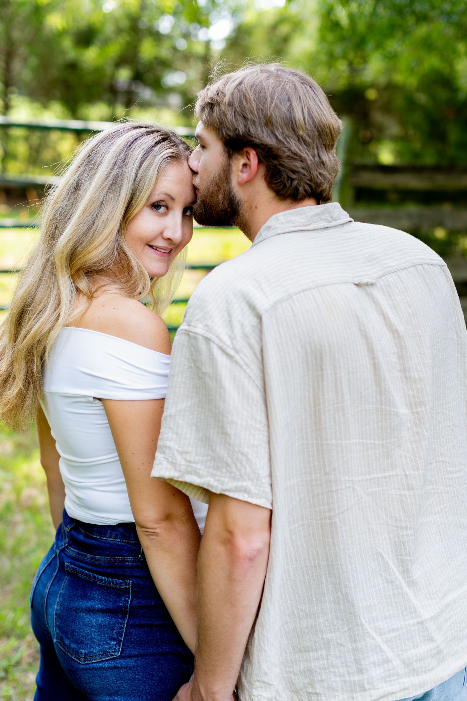 A couple shares a tender moment outdoors, man kissing woman's cheek as she smiles at camera in a lush green setting.