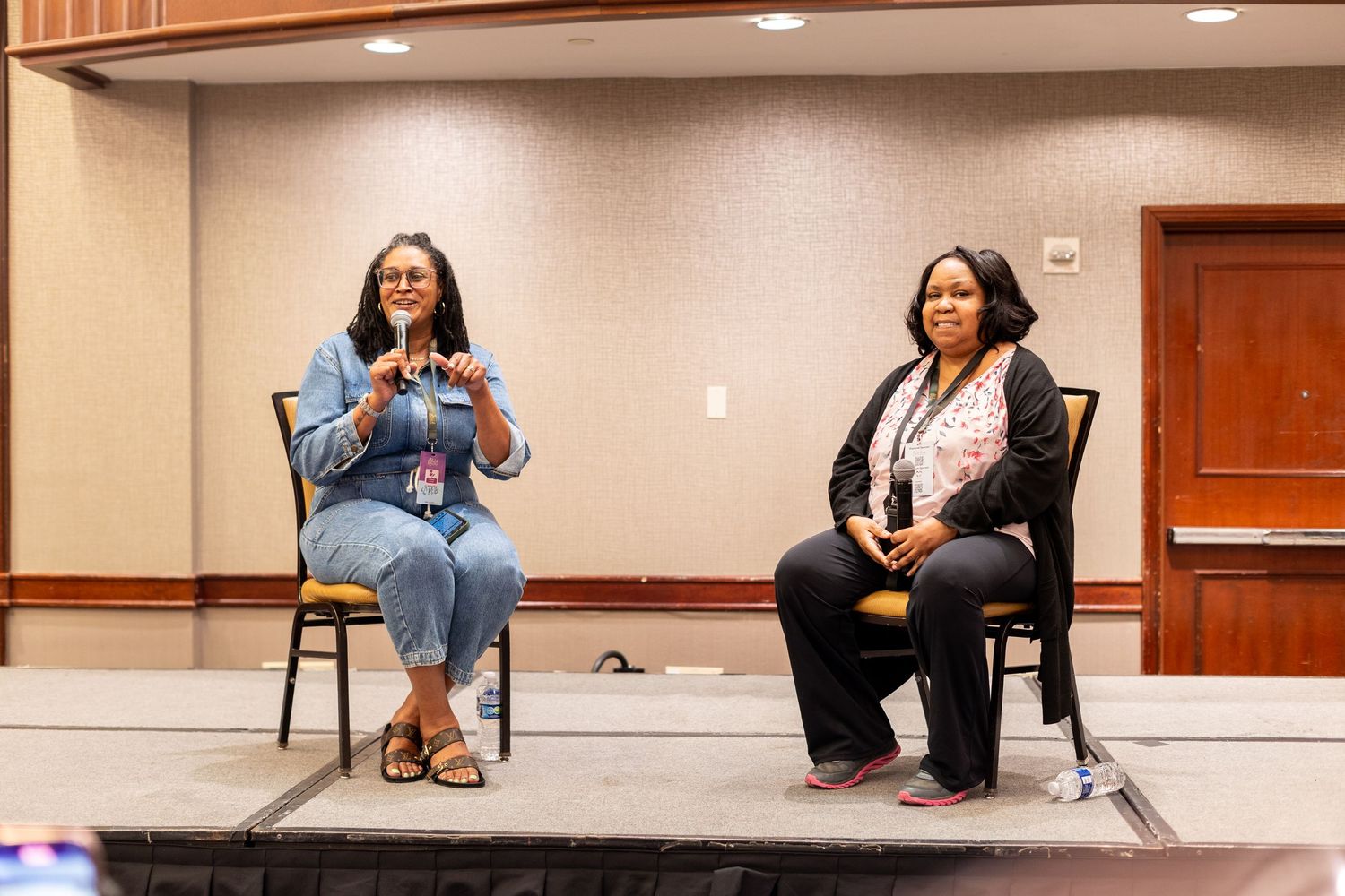 Two women seated on chairs on a stage during a panel discussion at an indoor conference event.