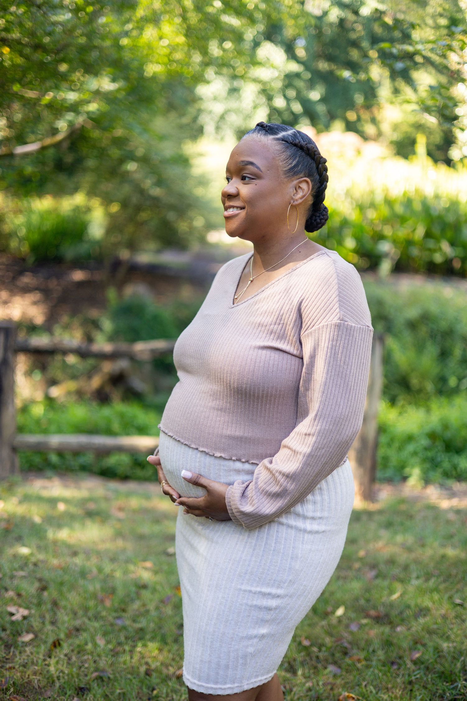 A pregnant woman in a light pink sweater and white skirt smiles while standing in a sunny garden setting.