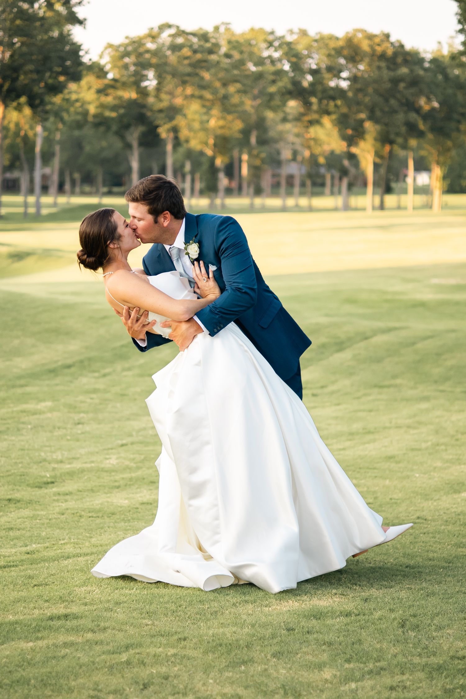 A romantic wedding couple shares a dip kiss on a sunlit golf course during their outdoor photo session.
