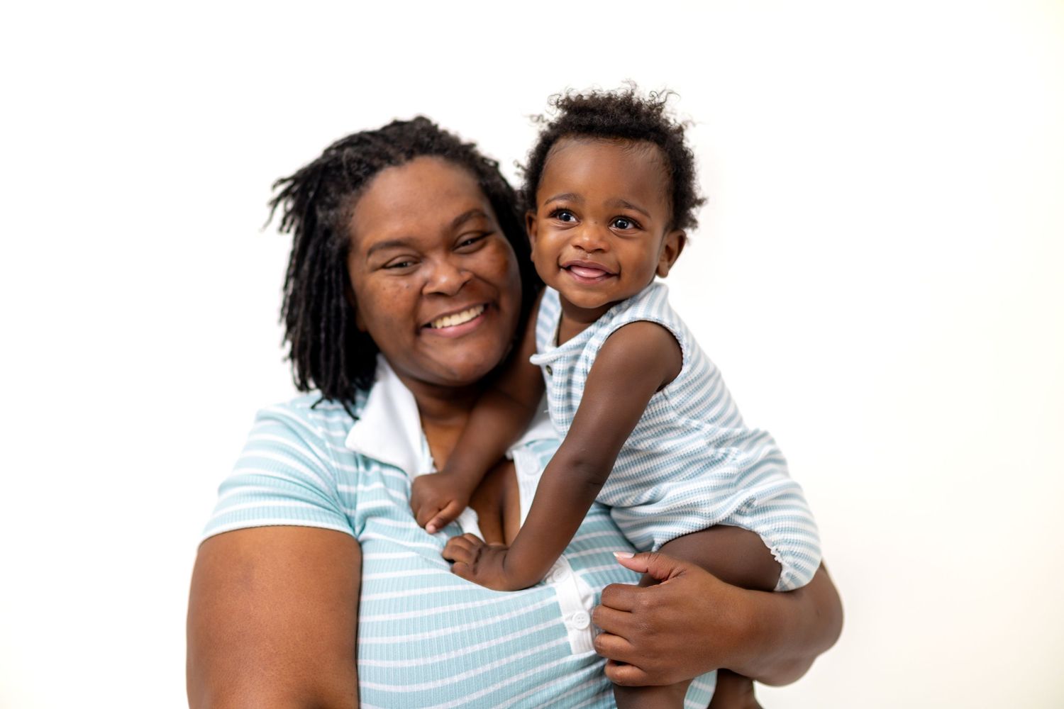 Smiling Black mother holding happy toddler in matching blue striped outfits against white background.