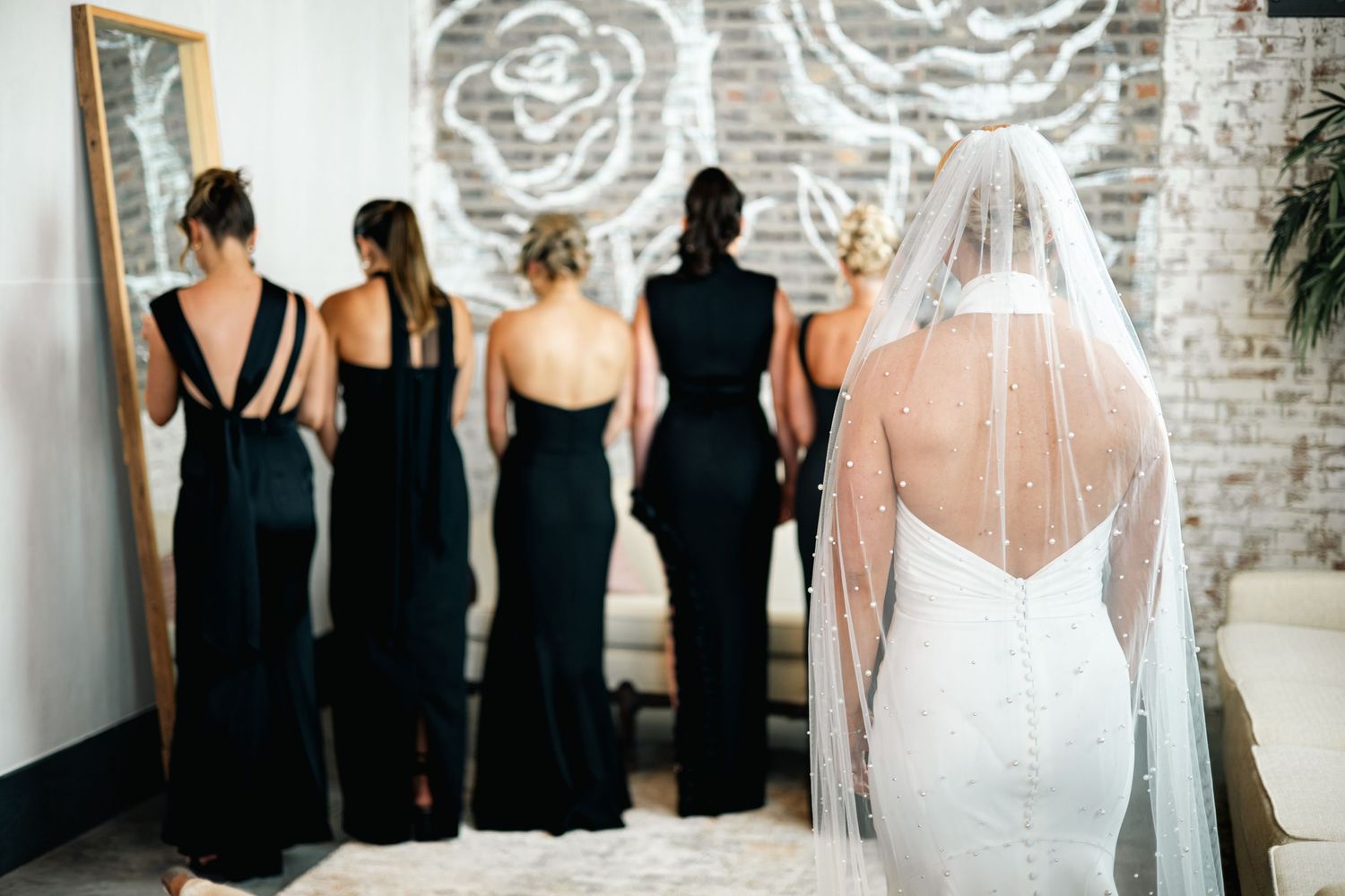 Wedding party lined up against white brick wall with bridesmaids in black dresses and bride in white gown.