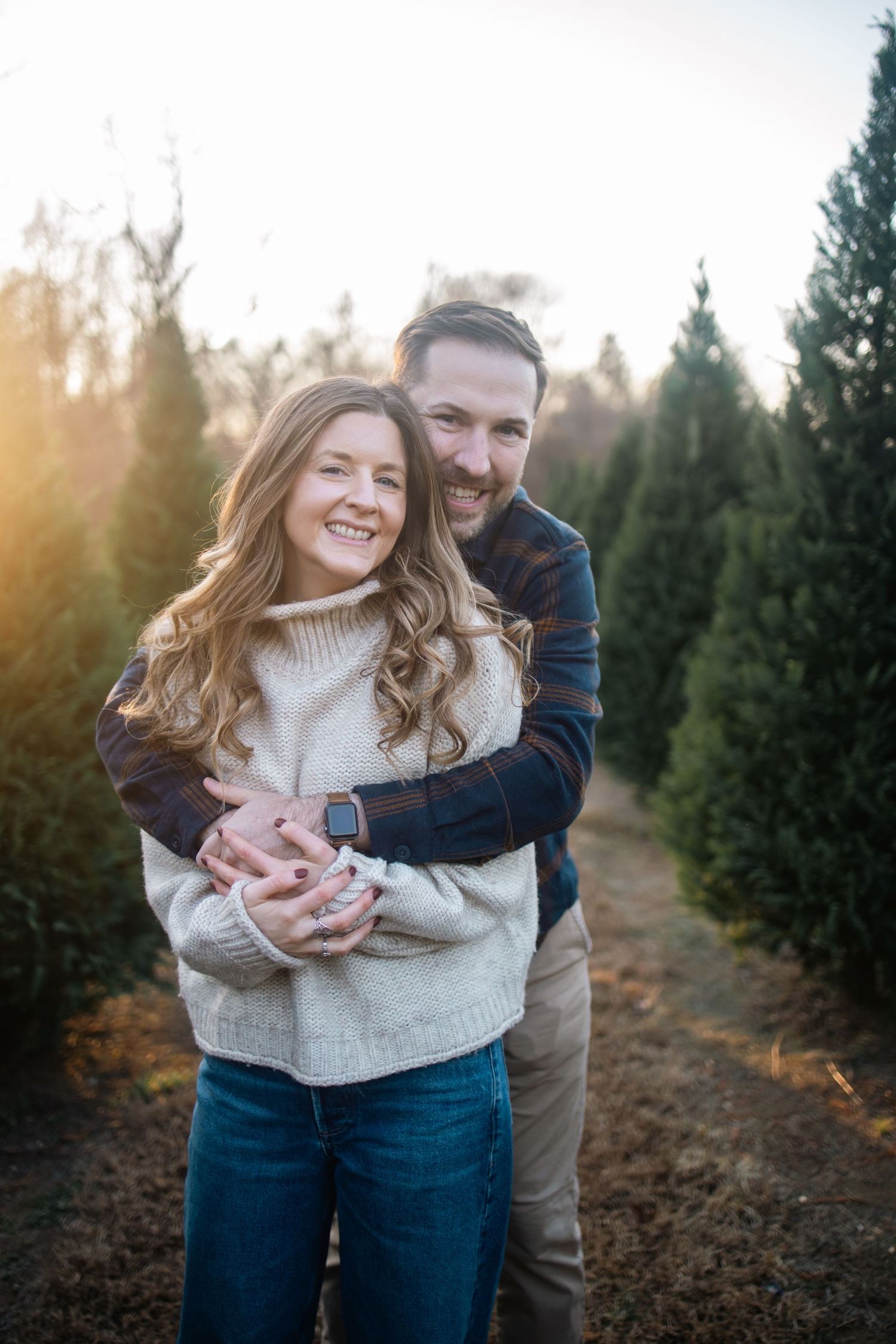 A couple embraces and laughs together while standing between rows of evergreen trees at a Christmas tree farm.