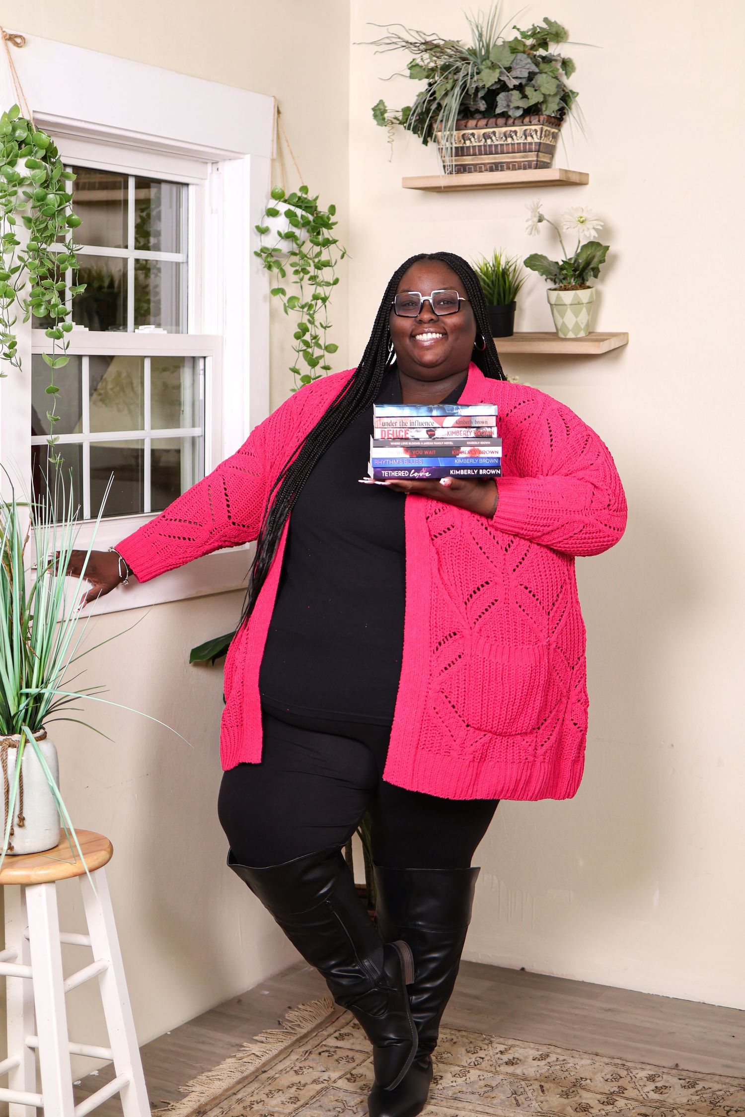 A person wearing a black top and pink cardigan poses in a bright interior space with plants and windows.