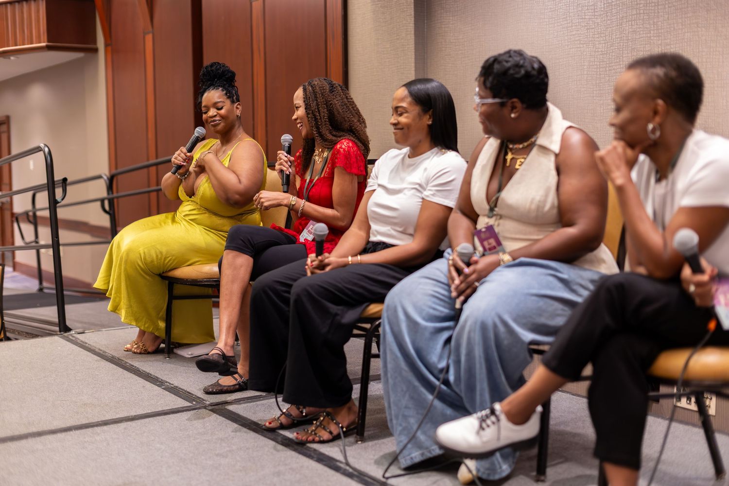 Six women seated in a row during a panel discussion, with two holding microphones in a modern conference room.