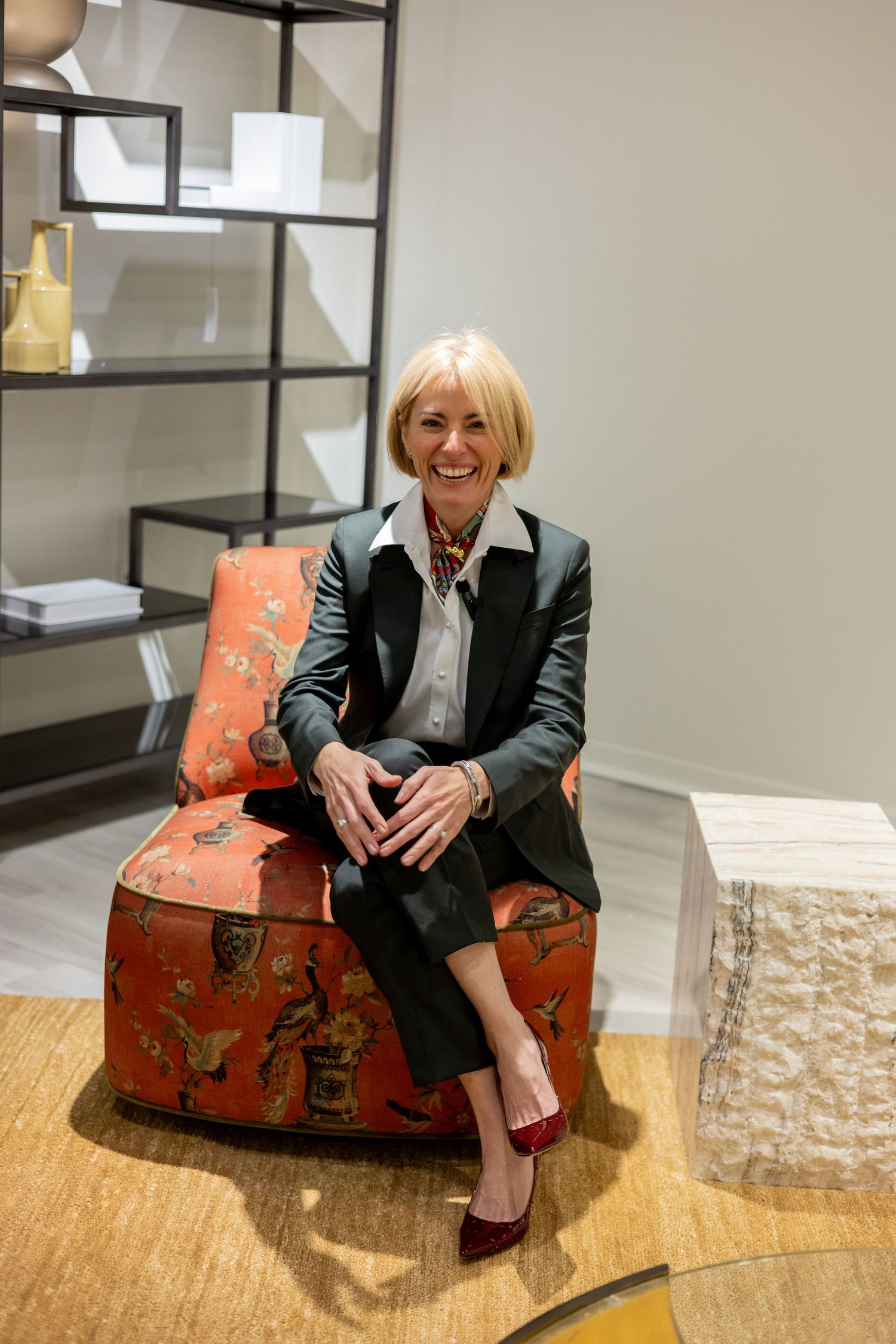 A person in a black blazer sits on a vintage red patterned suitcase in an interior space.