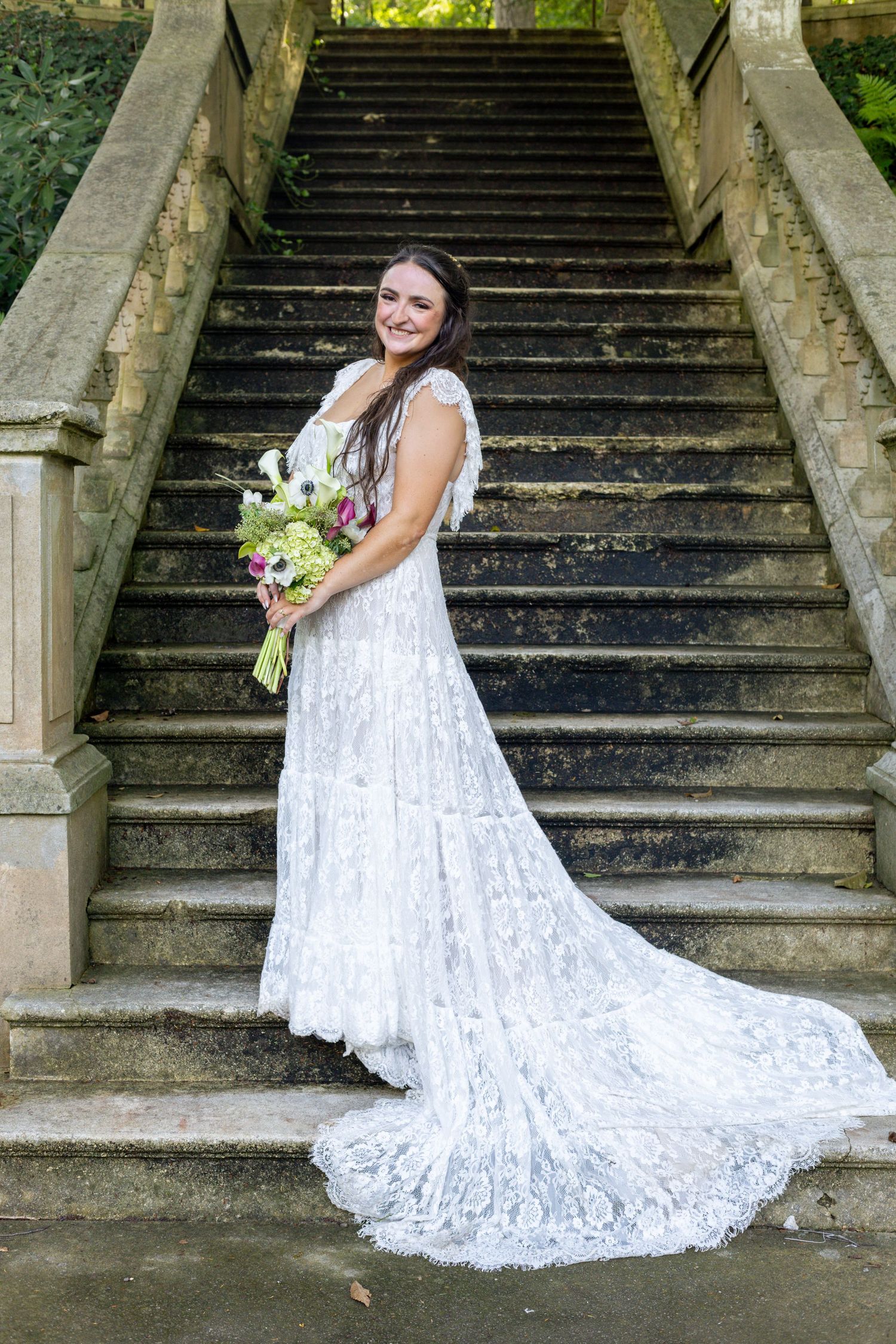 Bride in white lace gown holding floral bouquet poses on stone staircase outdoors.