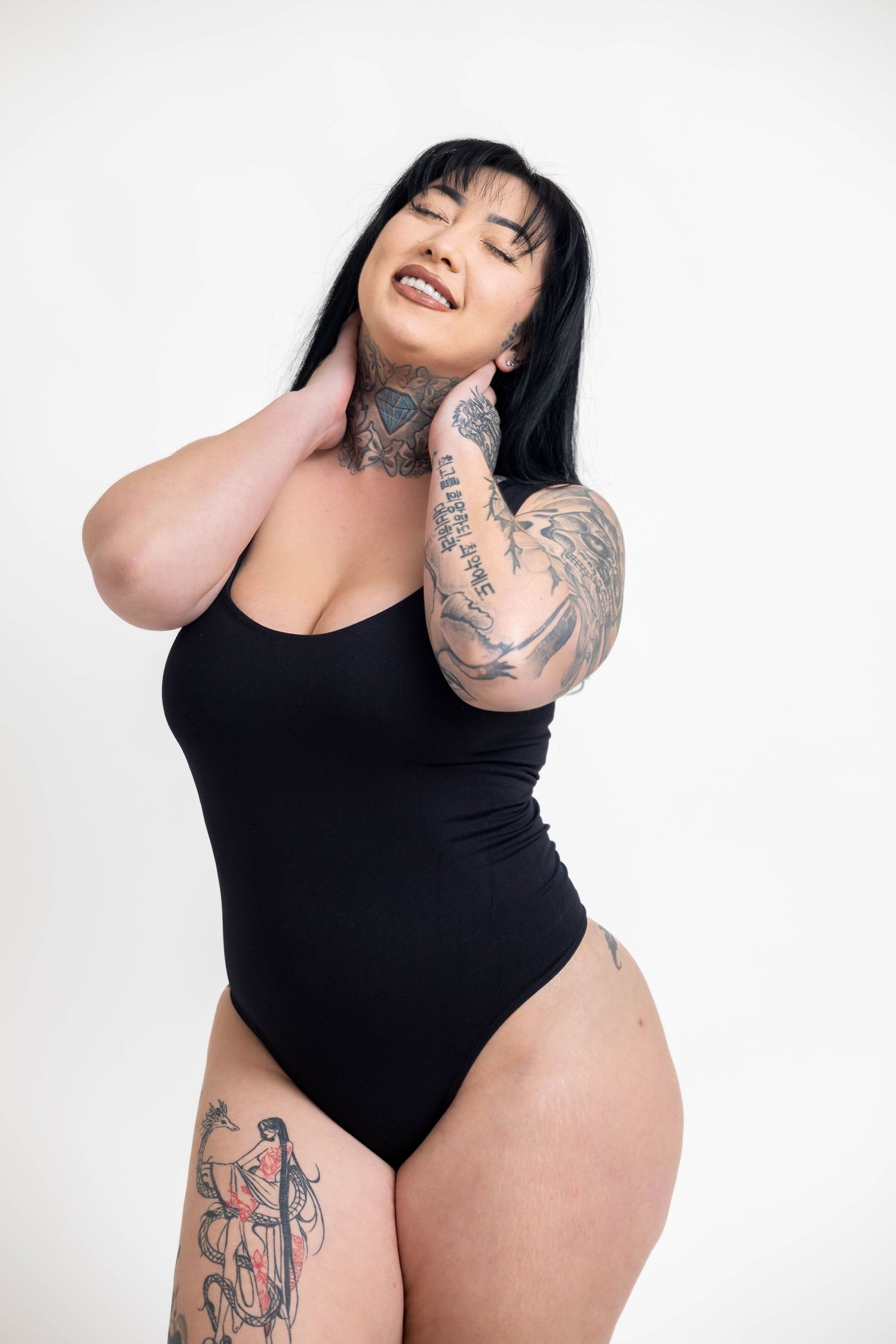 Model in black bodysuit poses confidently showing arm tattoos against white background.