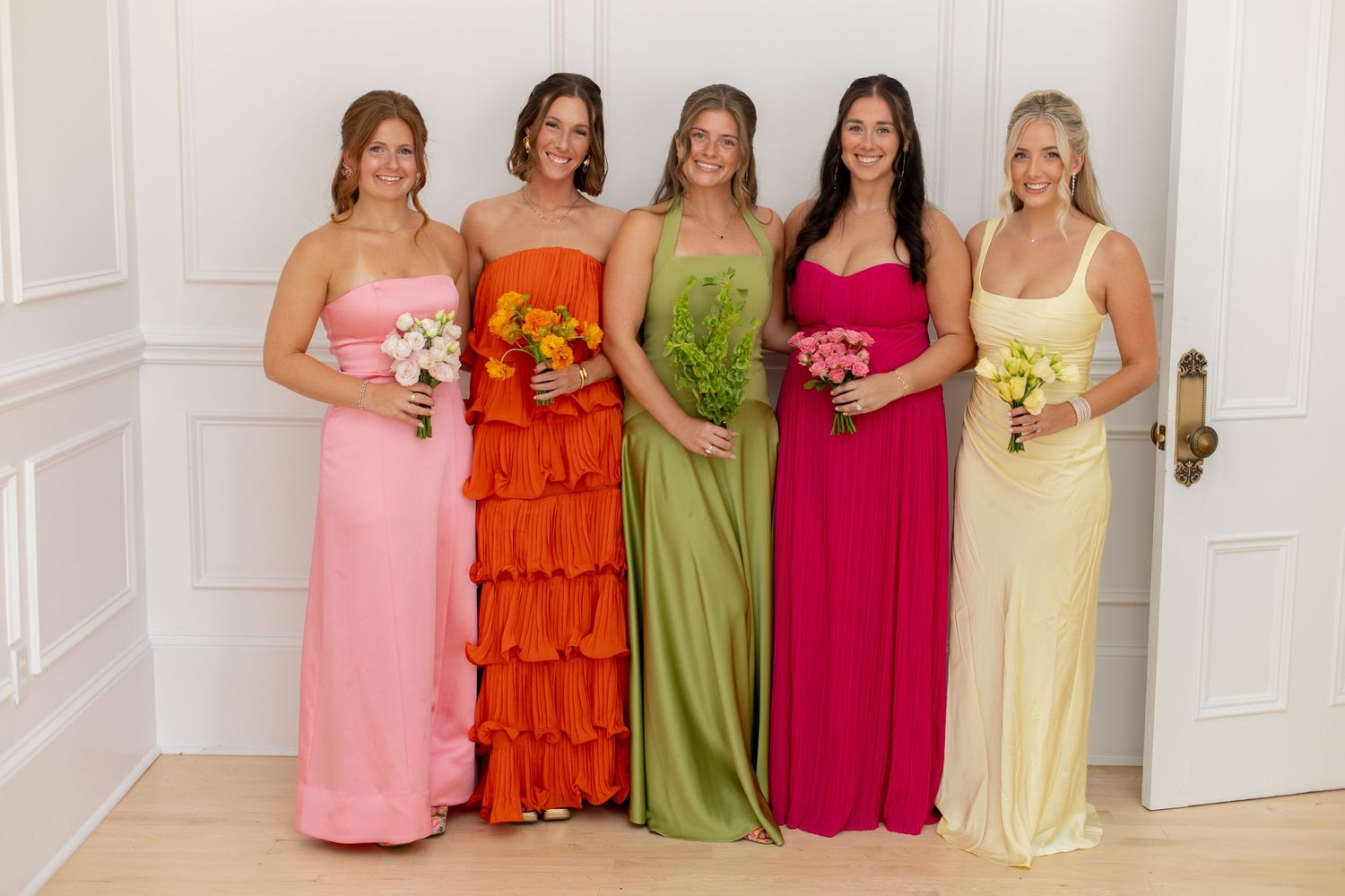 Five bridesmaids in colorful mismatched gowns holding bouquets, wearing pink, orange, green, and yellow dresses.