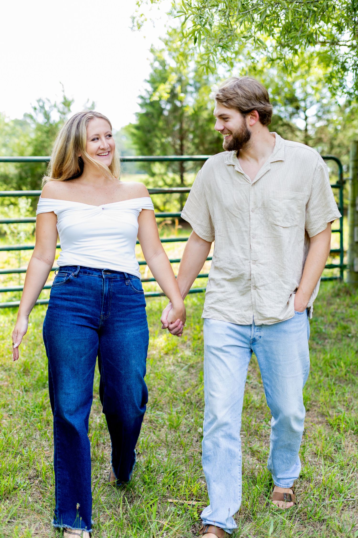 A smiling couple holds hands while walking outdoors near a metal gate surrounded by green trees.