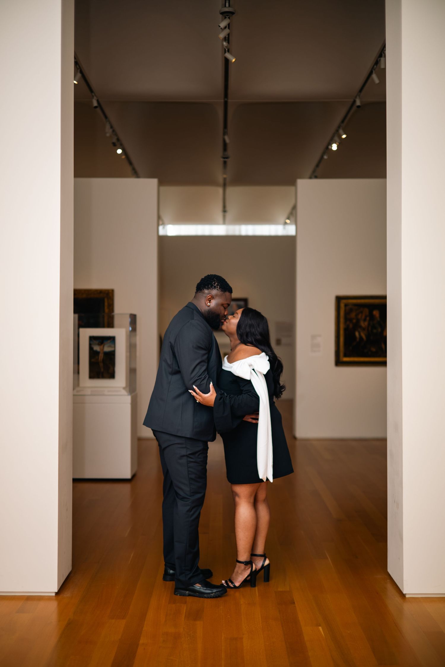 A romantic moment between two people in an art gallery with white walls and hardwood floors.