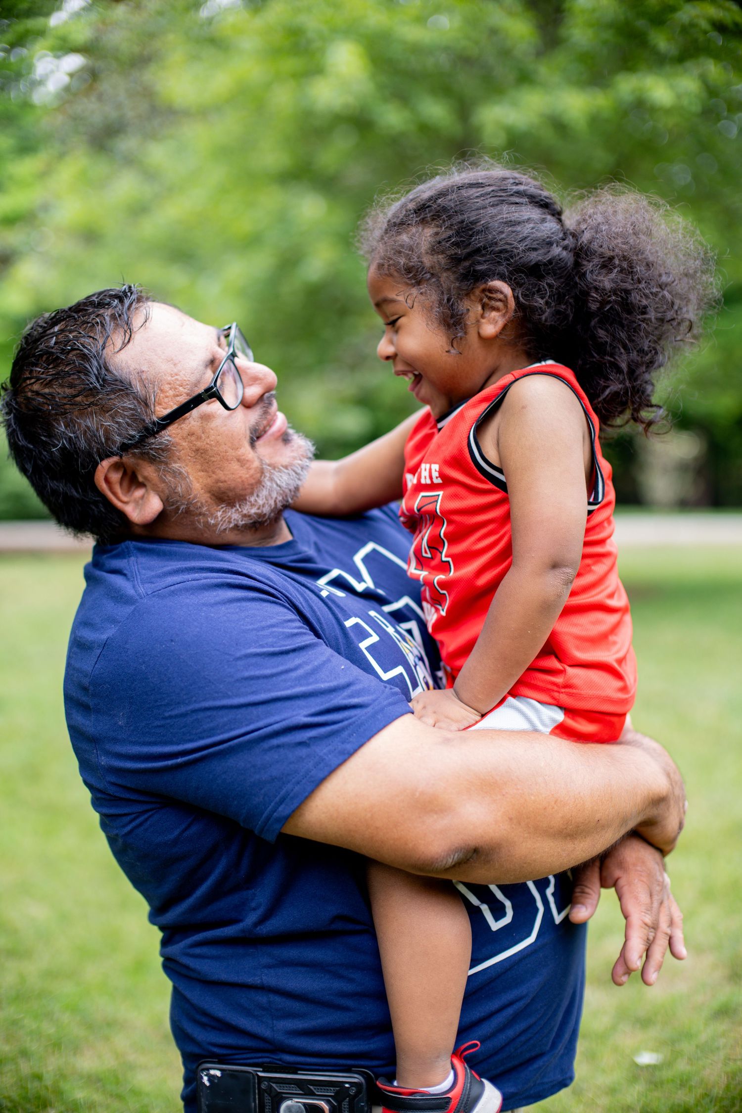 A man in a blue shirt holds a laughing young girl in a red jersey outdoors in a green park.