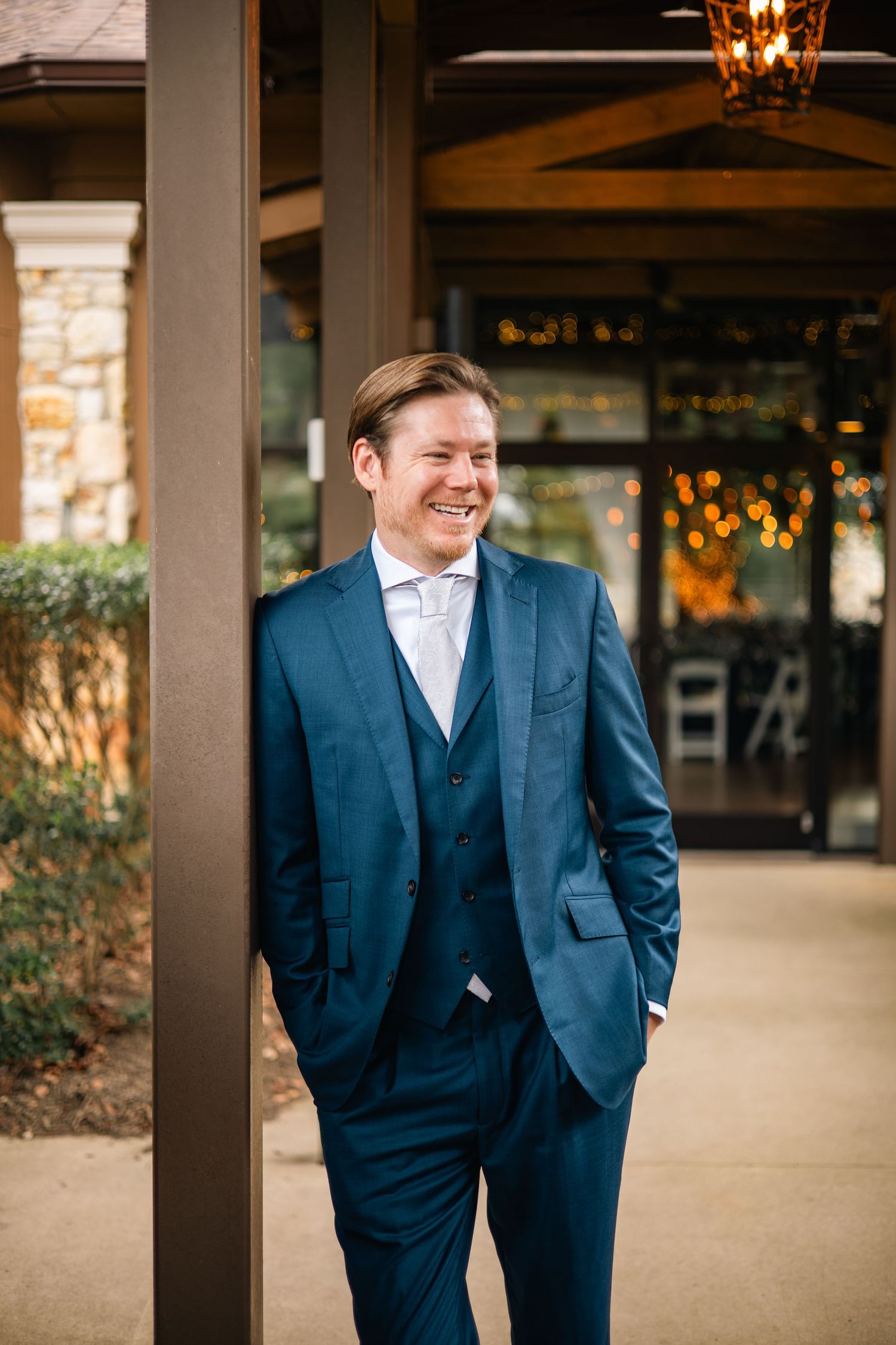 A person wearing a navy blue three-piece suit stands under covered walkway columns at an elegant venue.