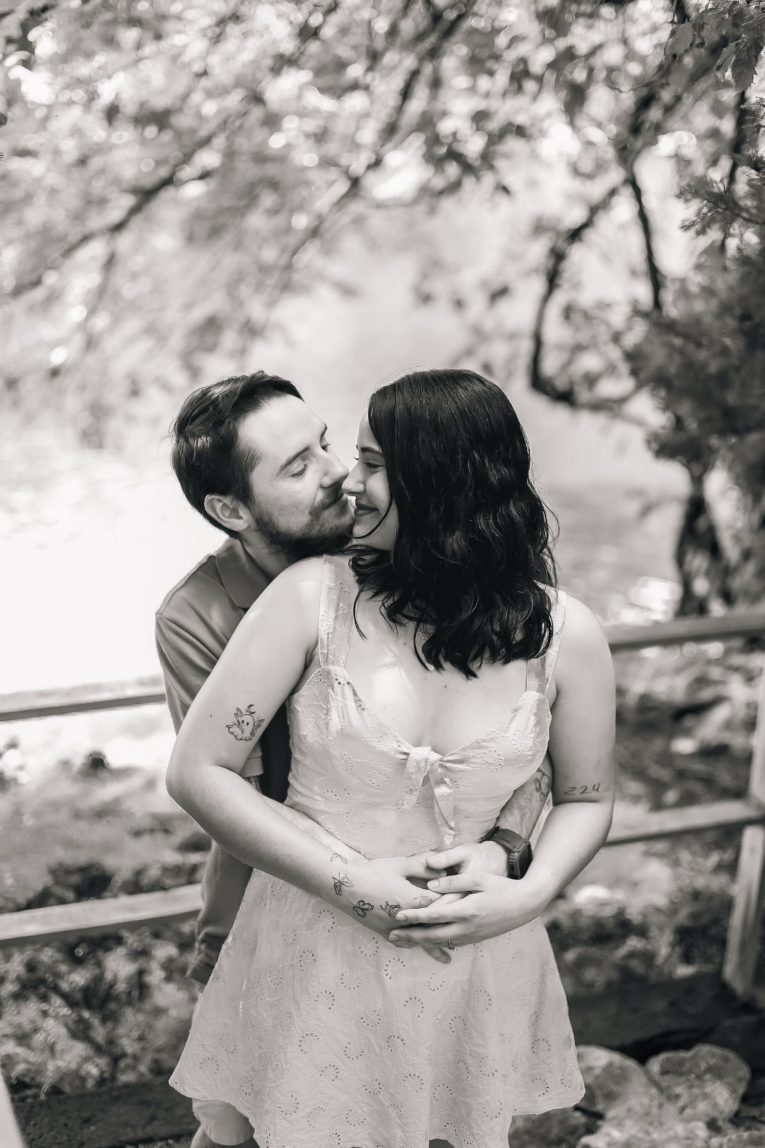 A romantic black and white portrait of a couple embracing outdoors with trees and natural scenery in the background.