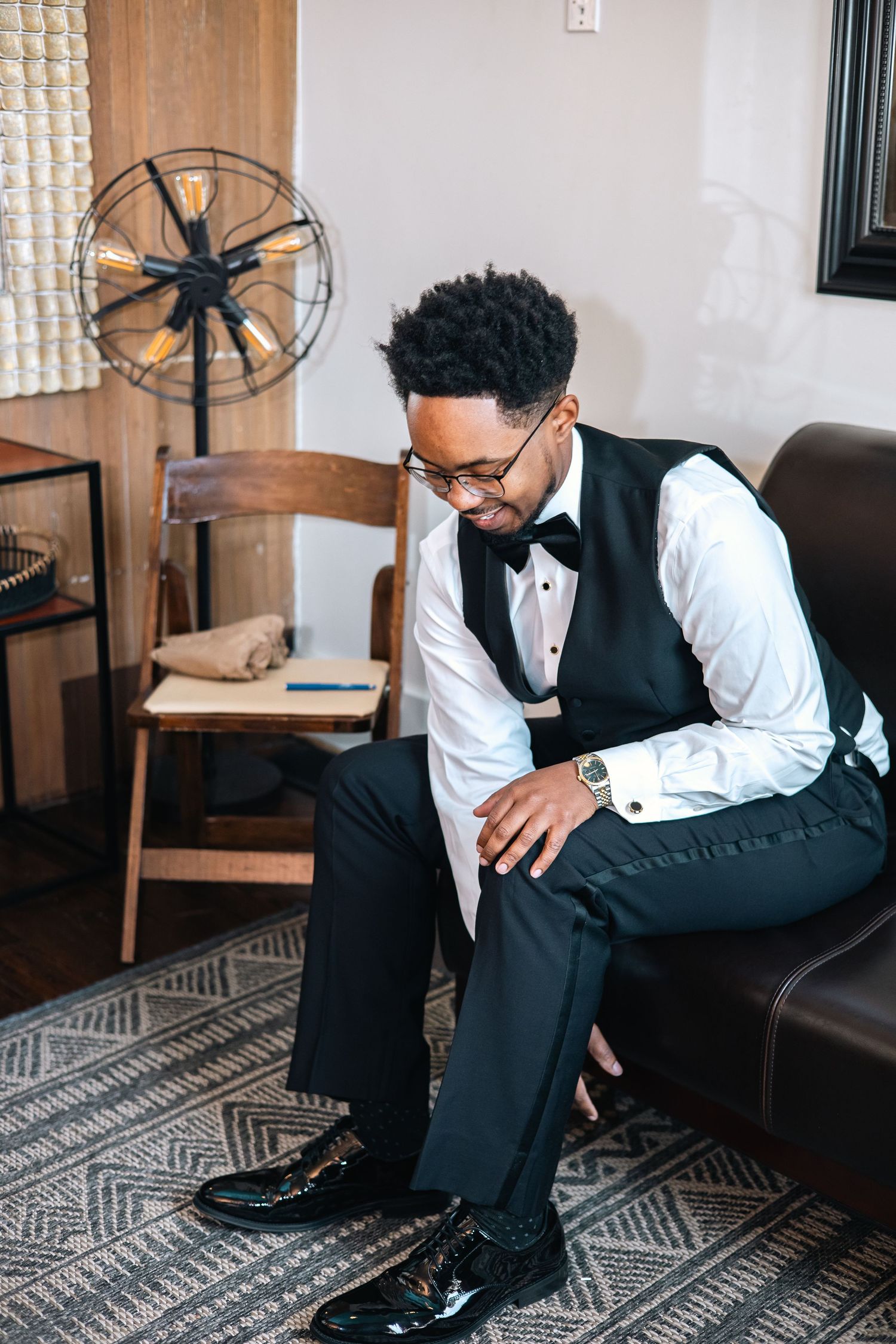 Professional in formal attire sits thoughtfully on brown leather couch with geometric patterned rug in stylish room setting.