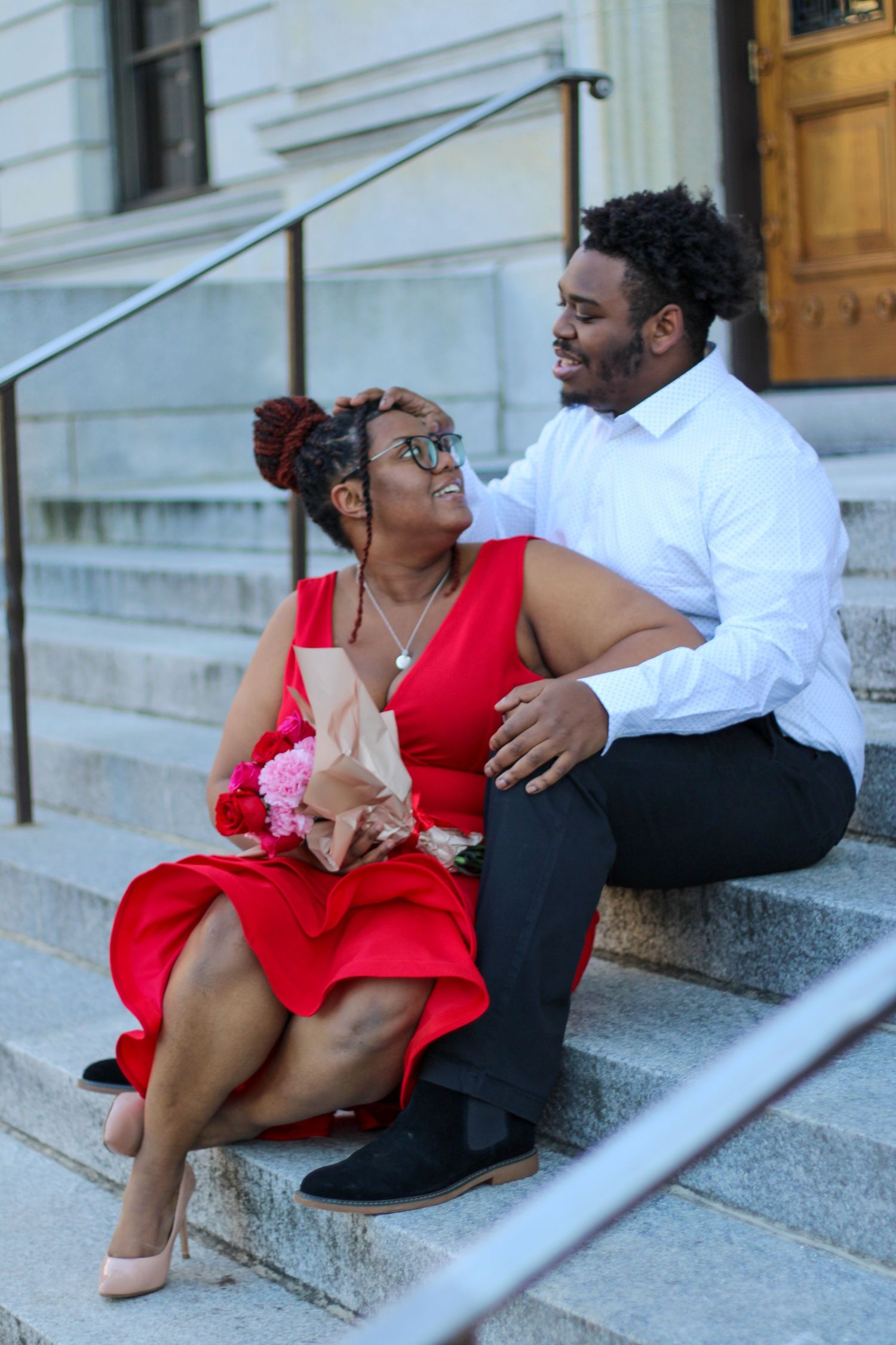 A couple sits together on stone steps, with one in a red dress and other in white shirt.